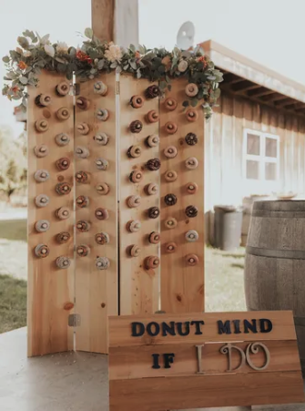 Wooden display available for rent with numerous small cans attached in rows, decorated with flowers on top, and a sign that reads "Donut Mind If I Do" in front.