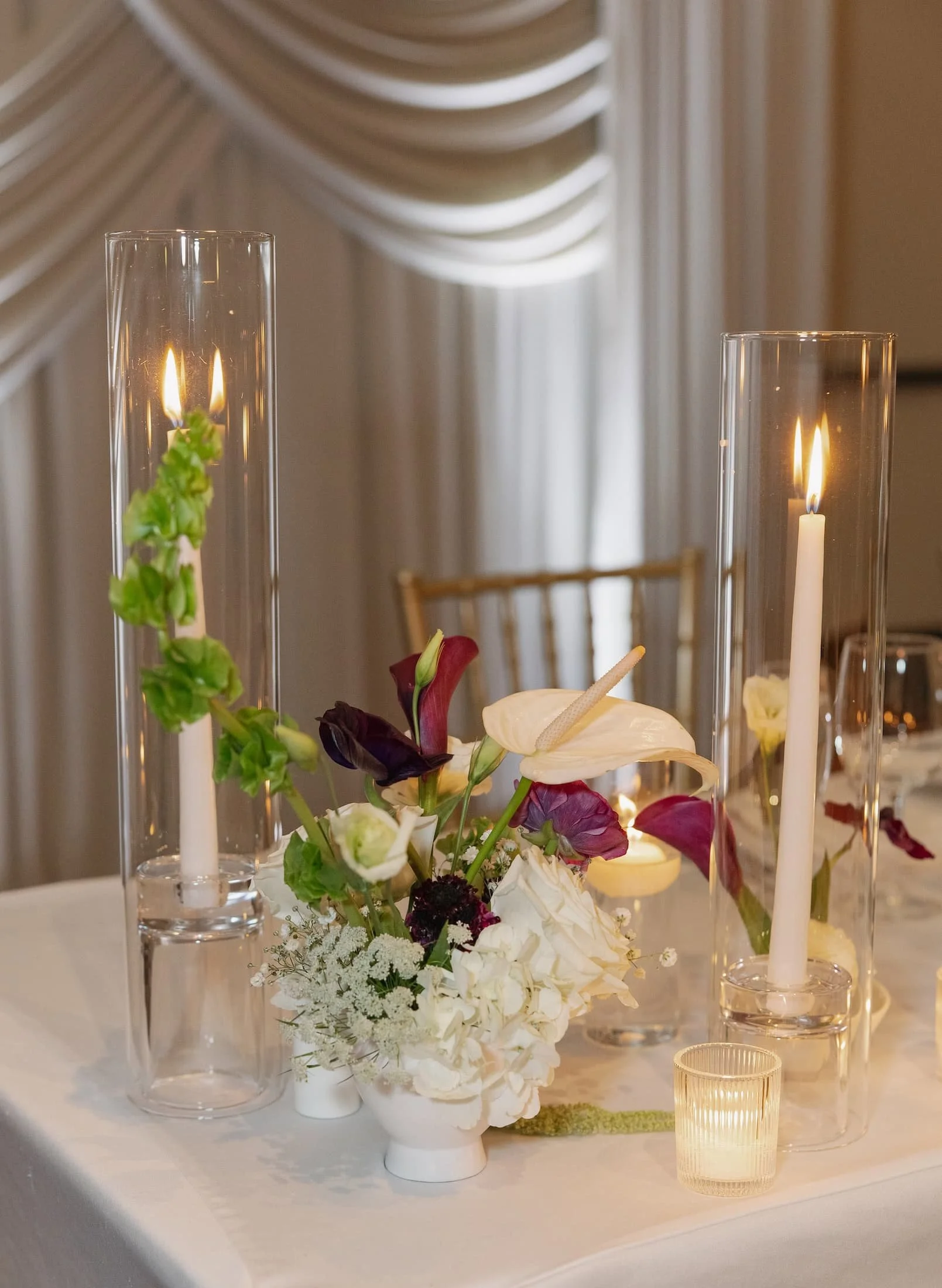 Elegant table centerpiece with white and dark red flowers, surrounded by tall glass candle holders with lit white candles, and a small votive candle, on a white tablecloth at the Davenport Hotel in Spokane, Washington