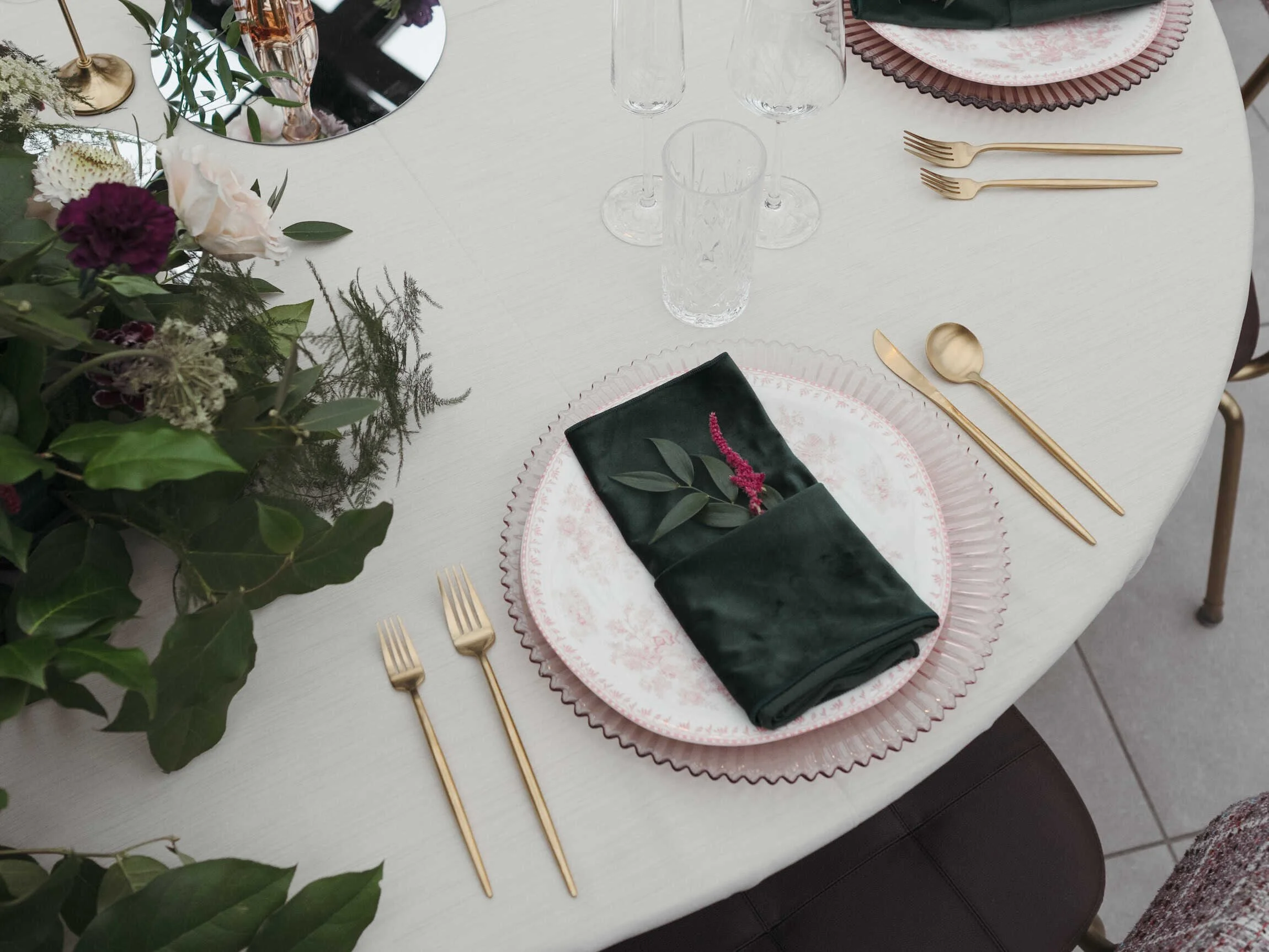 Elegant table setting with pink and white patterned plates, gold utensils, a dark green napkin with greenery, and a floral centerpiece on a white round table.