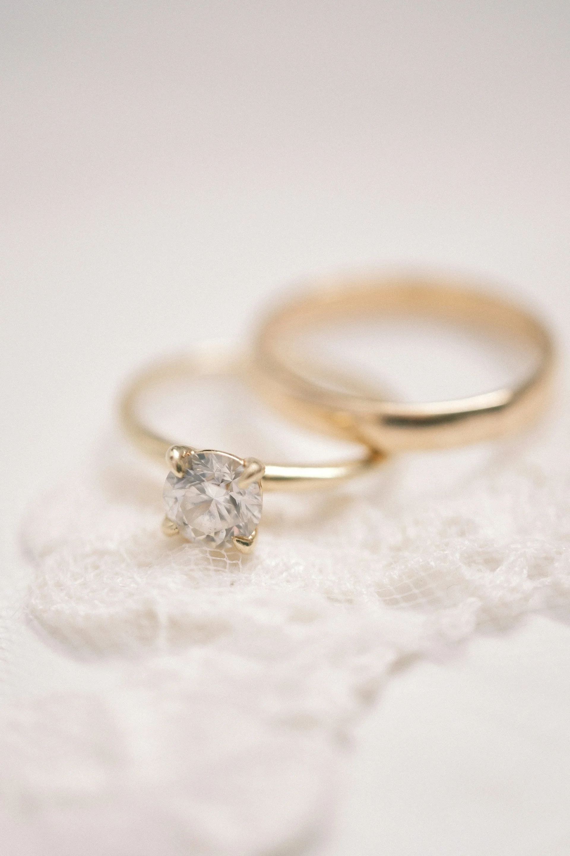 Close-up of a diamond engagement ring with a gold band and a single large diamond, placed next to a plain gold wedding band on white lace fabric at the Davenport Hotel in Spokane, Washington