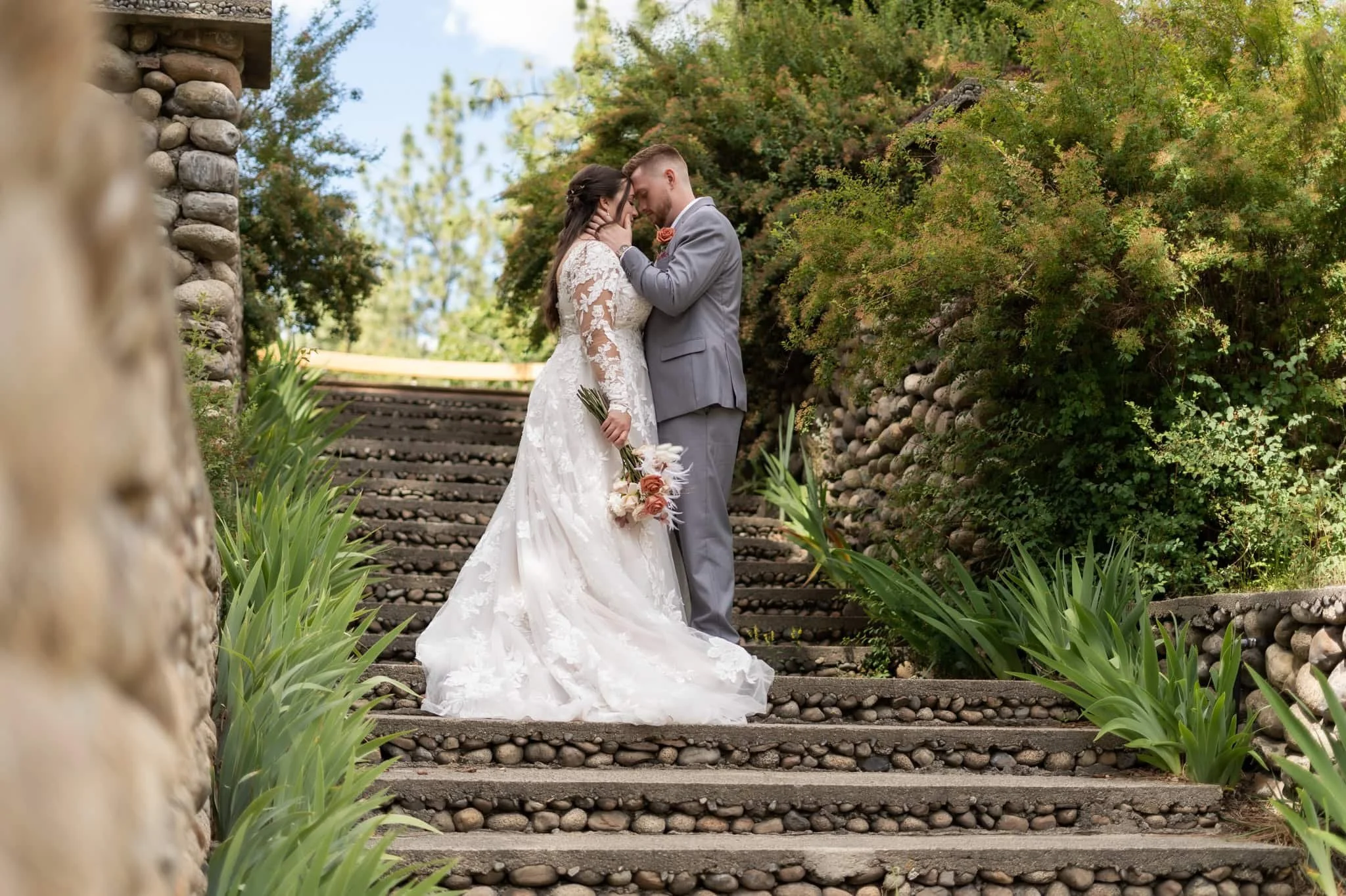 A bride and groom sharing a tender moment on outdoor stone stairs lined with green plants and surrounded by trees in Spokane