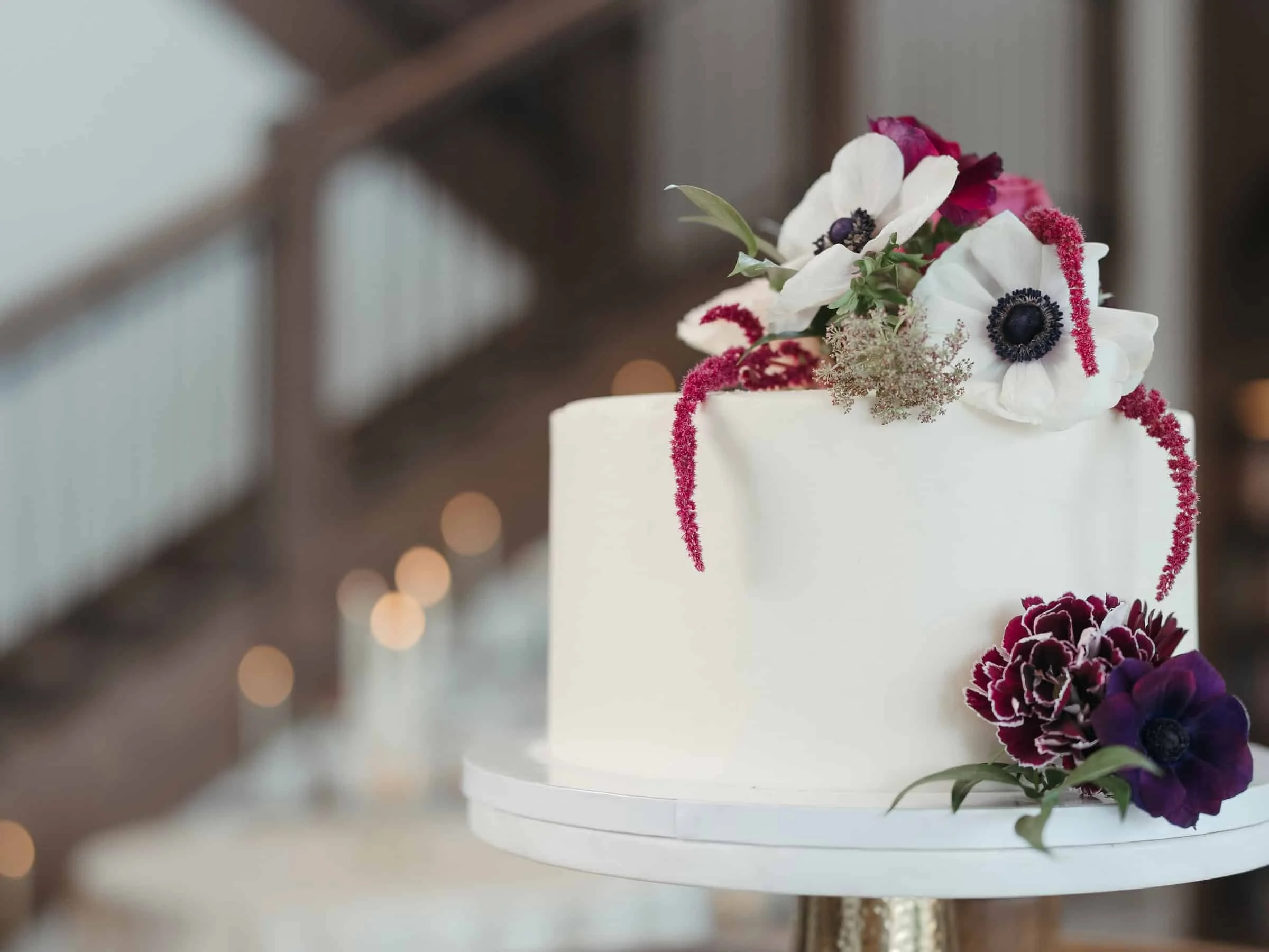 White wedding cake topped with white, purple, and pink flowers, placed on a gold stand at Kindred & Co in Post Falls, Idaho