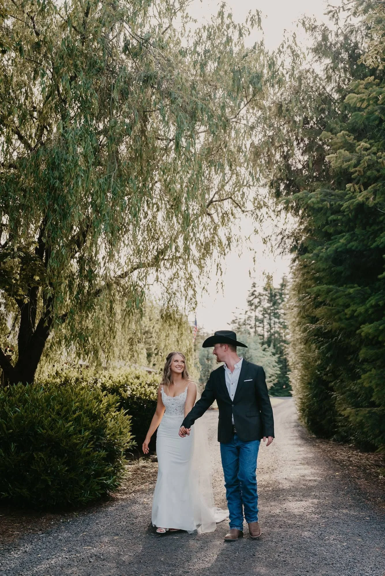 A couple in wedding attire walking hand in hand down a dirt path surrounded by lush green trees and bushes, with the woman wearing a white wedding dress and the man wearing a dark suit, white shirt, jeans, and a cowboy hat in Spokane