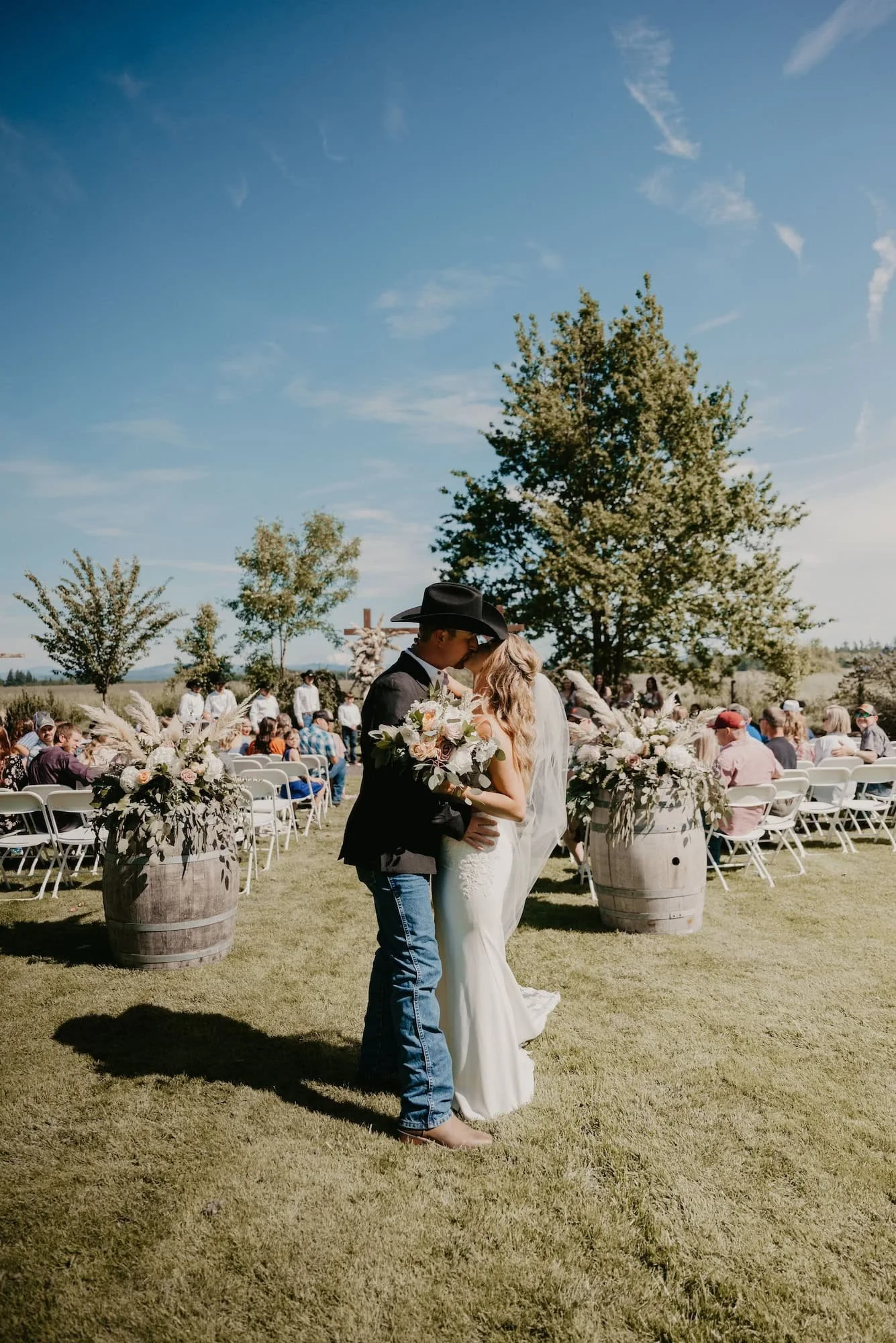 A couple getting married outdoors, kissing, with guests seated in the background and floral arrangements in large barrels in Spokane