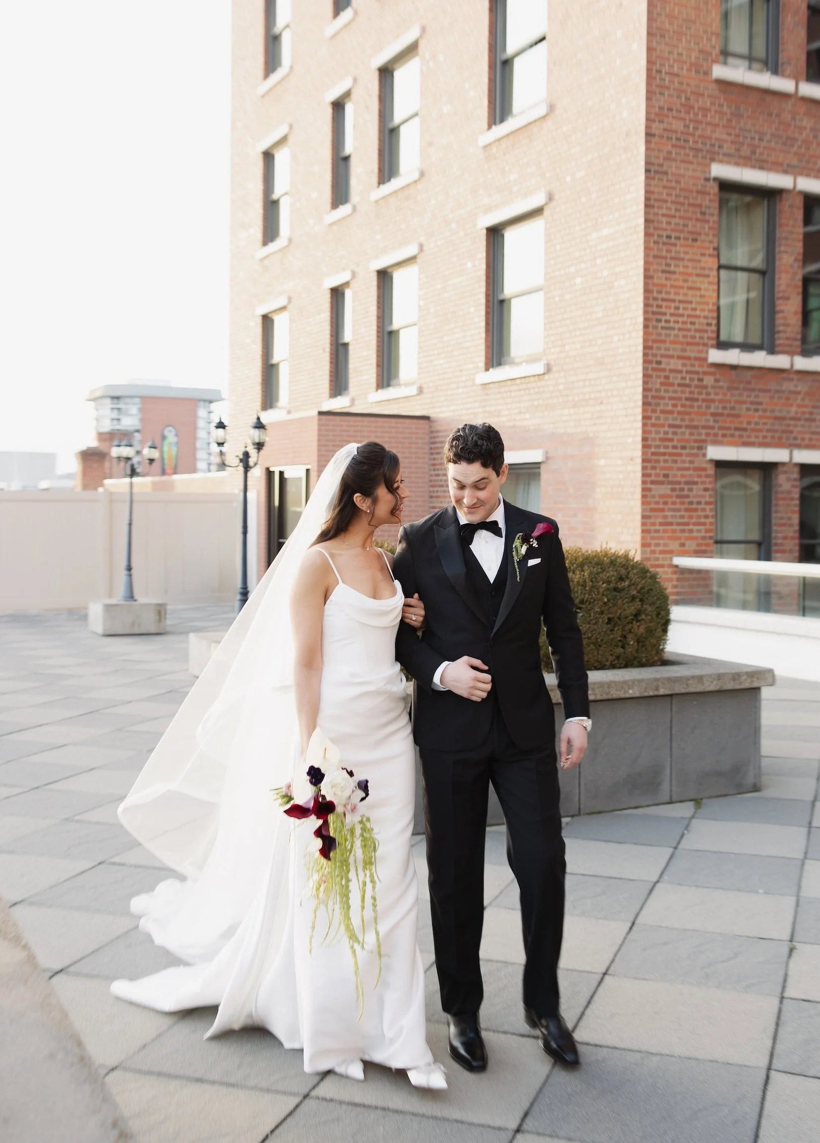 Bride and groom walking together outside on a city street in Spokane, smiling. The bride wears a white wedding gown and veil, holding a bouquet. The groom wears a black tuxedo with a bow tie and boutonniere.