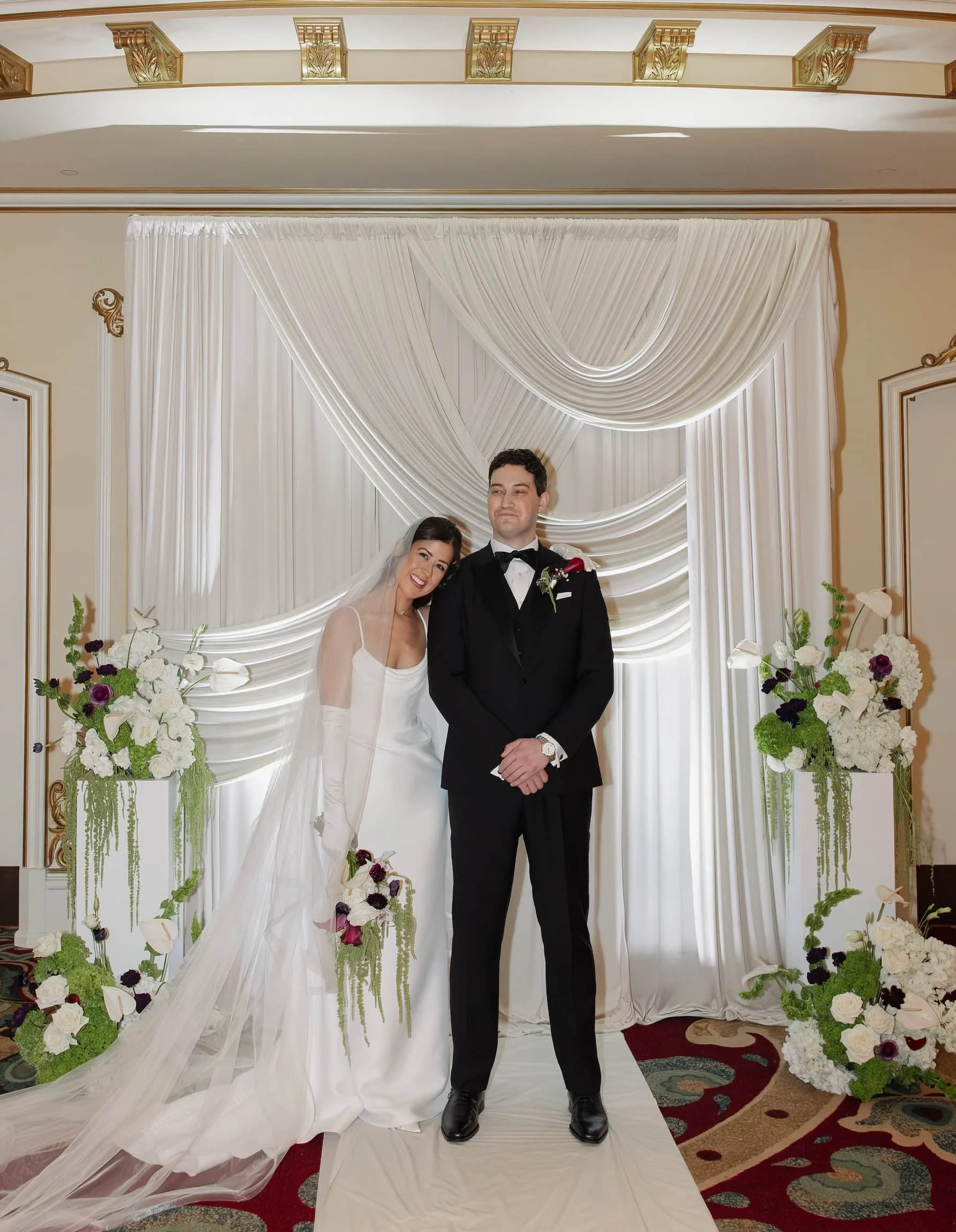 A bride and groom standing together during their wedding ceremony in front of a white draped backdrop with floral arrangements at the Davenport Hotel.