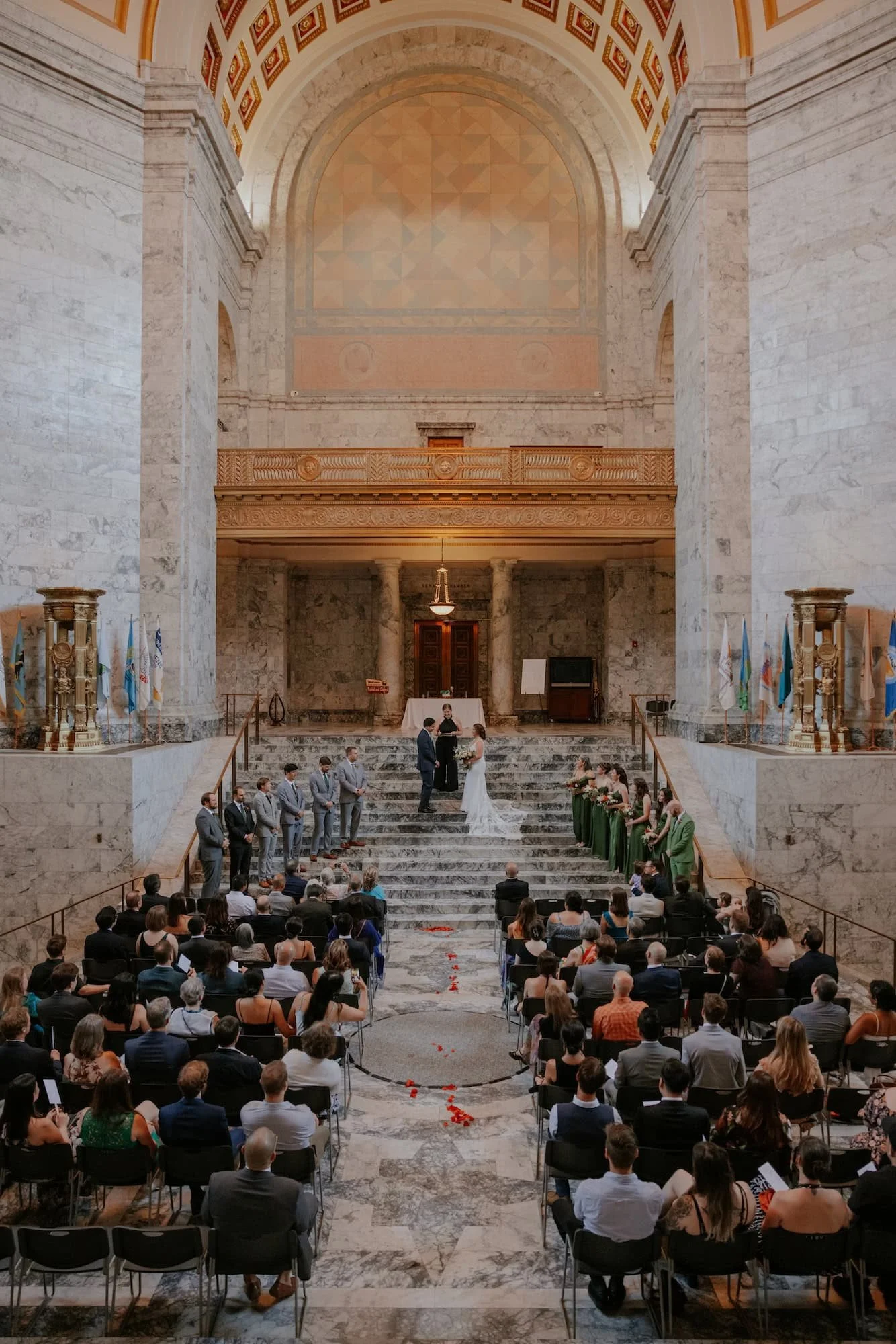 A wedding ceremony taking place inside a grand, marble hall with high ceilings and a large arched wall at the altar. The bride and groom stand on the steps facing each other, with the officiant behind them. Bridesmaids in green dresses stand to the right, and groomsmen in gray suits to the left. Guests are seated facing the altar, with some standing. Rose petals are scattered along the aisle.