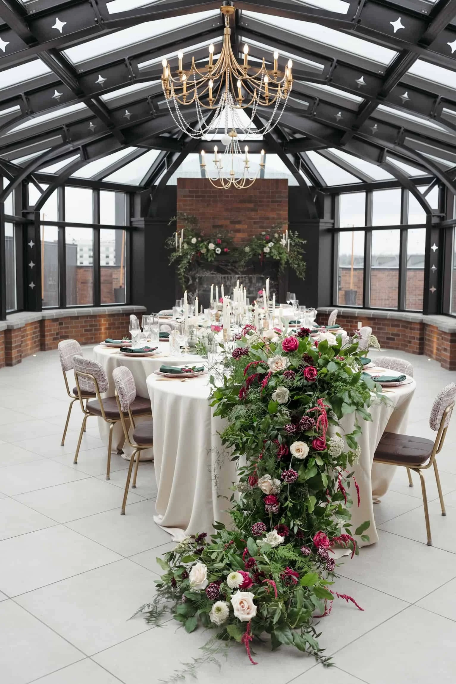 Elegant indoor wedding reception with a long table decorated with pink and white flowers, surrounded by chairs, and a brick fireplace with floral arrangements and candles in the background. The space has a glass ceiling with black framing and a large chandelier hanging above.