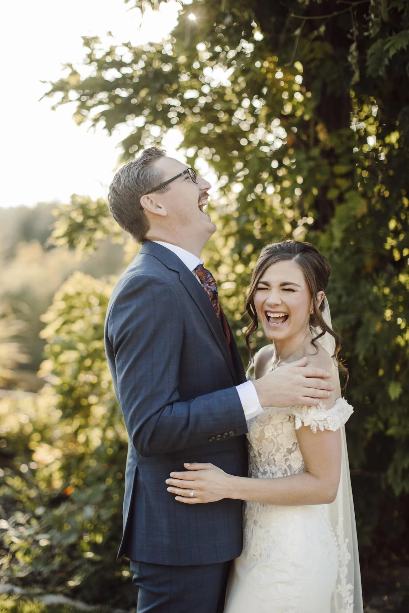 A newlywed couple laughing together outdoors with sunlight filtering through trees in the background in Spokane