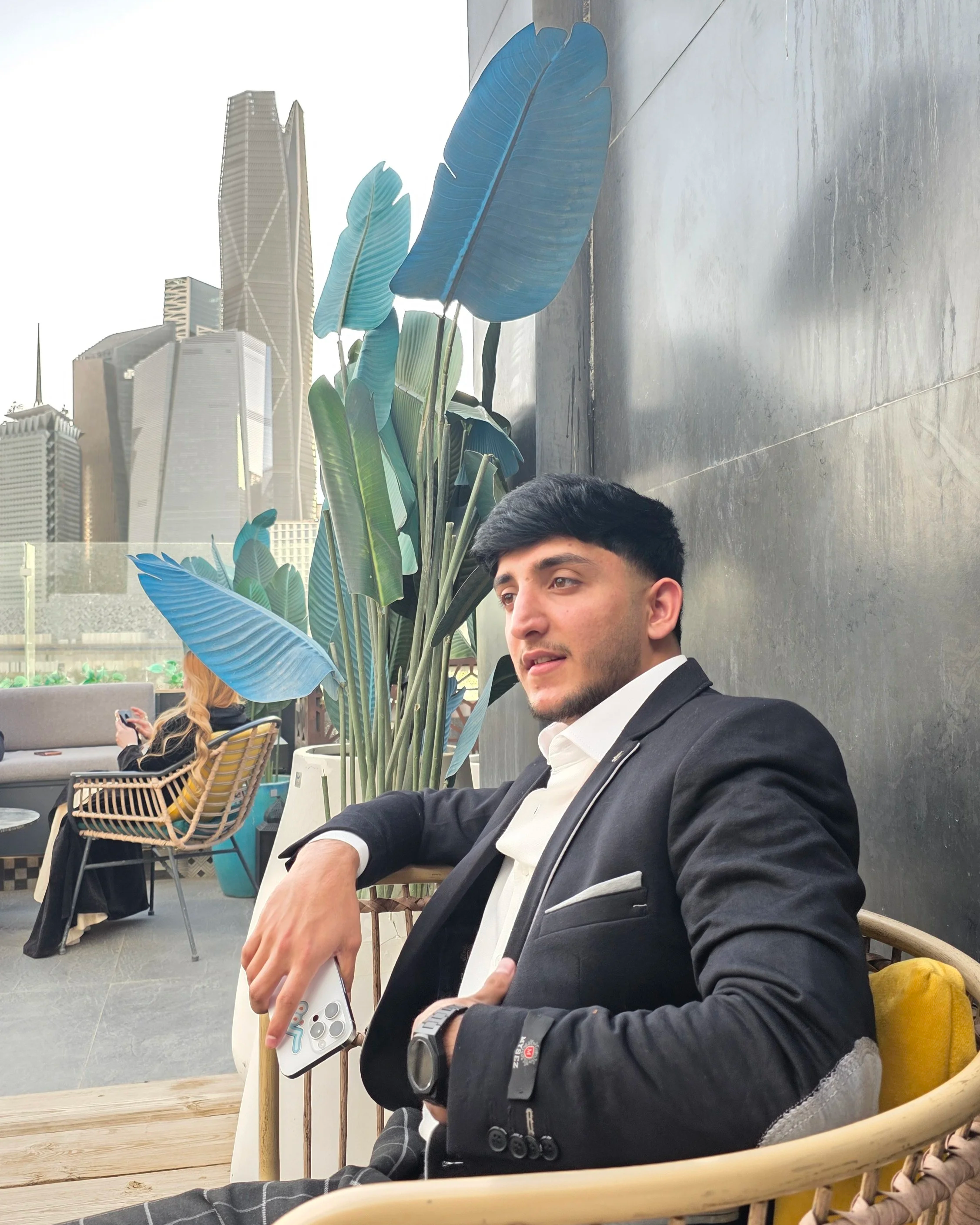 A man in a black blazer and white shirt sitting outdoors on a patio with large blue-green plants and tall buildings in the background.