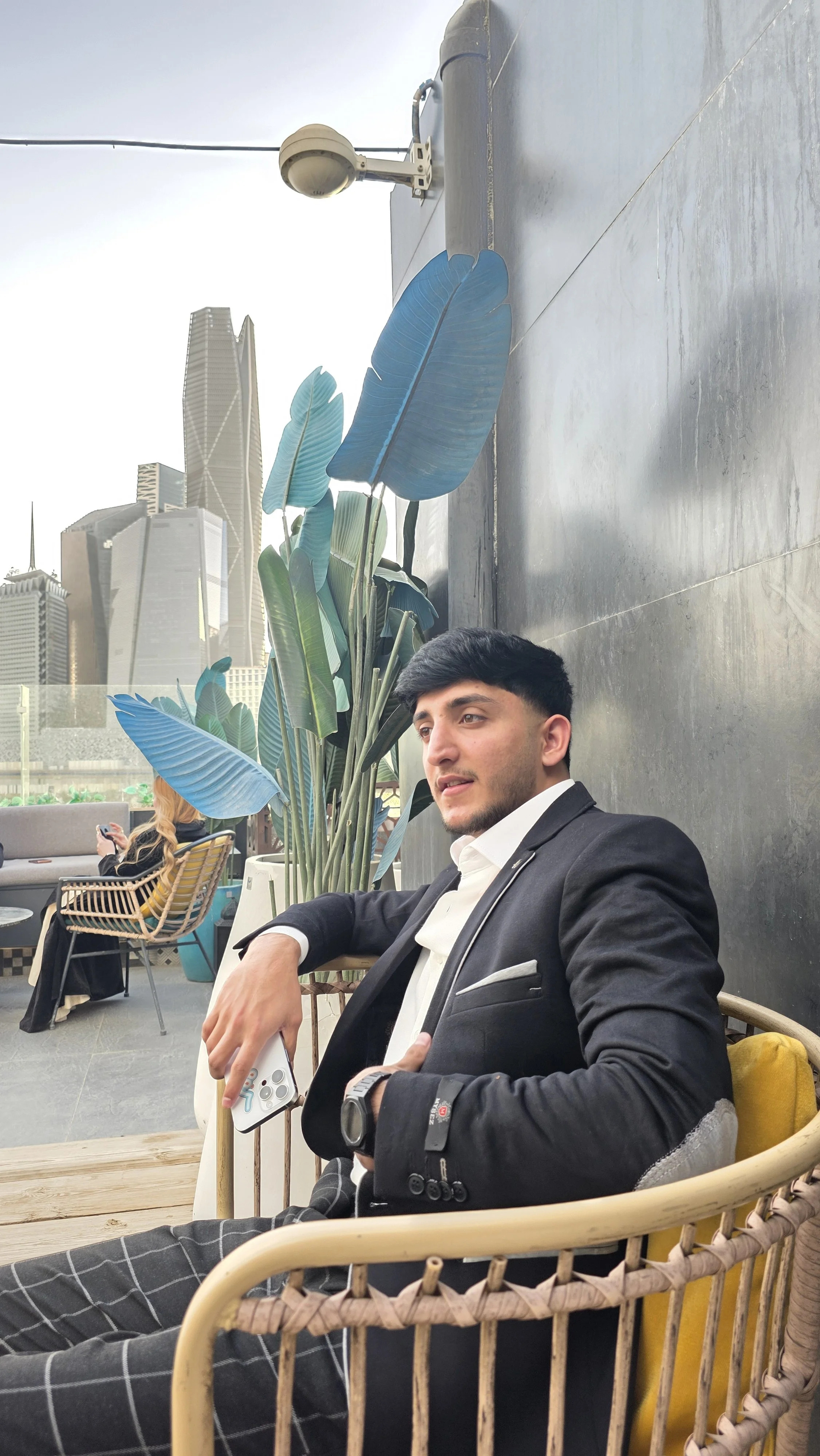 A man in a black suit and white shirt sitting on a chair outdoors, holding a smartphone with a city skyline in the background, and a large green plant with blue leaves nearby.