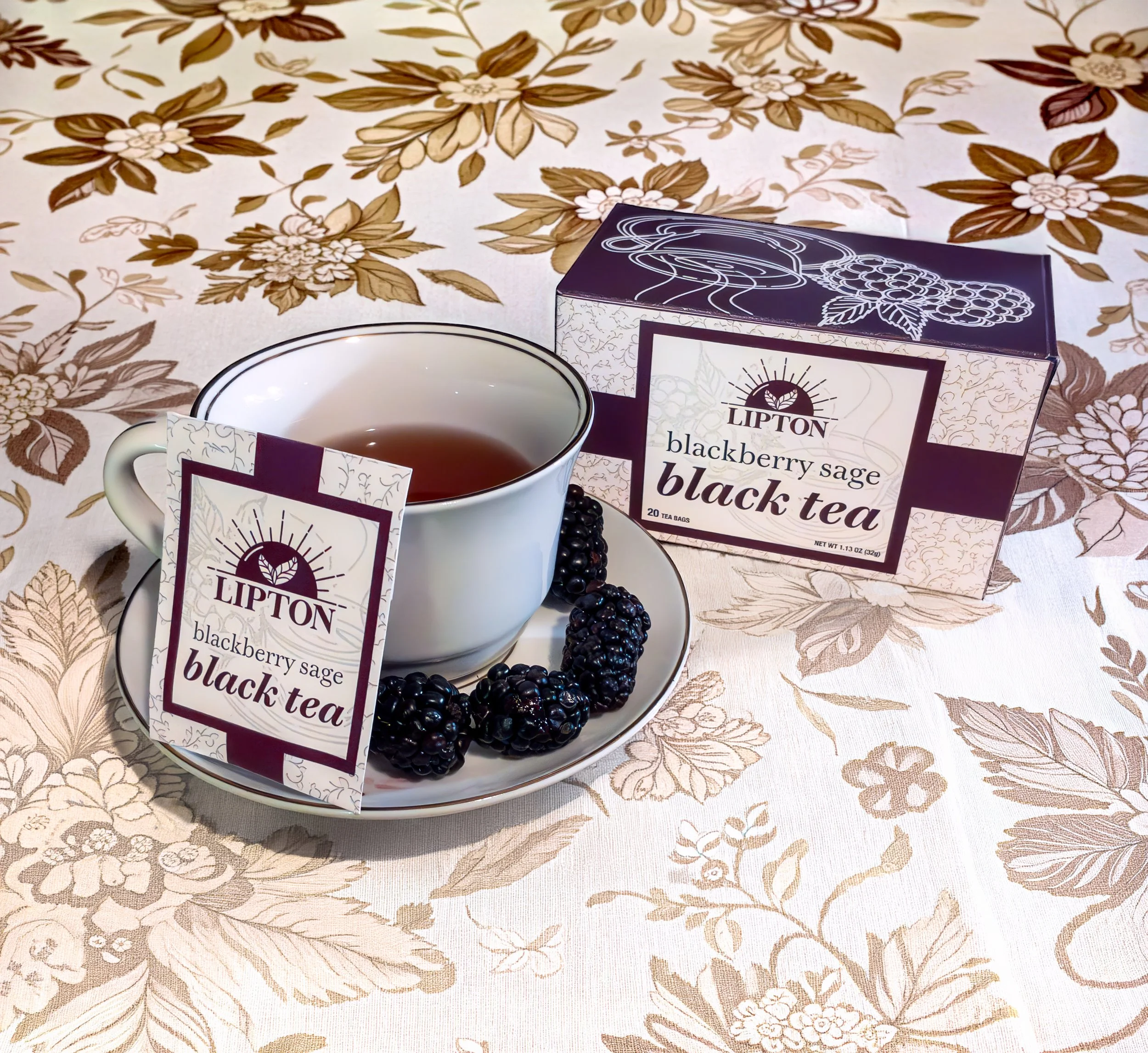 A cup of blackberry sage black tea on a saucer decorated with blackberries, with a box and packet of Lipton blackberry sage black tea on a floral tablecloth.