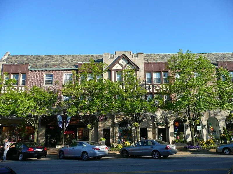 A street view of a multi-story building with shops on the ground floor, trees in front, parked cars along the street, and a clear blue sky.