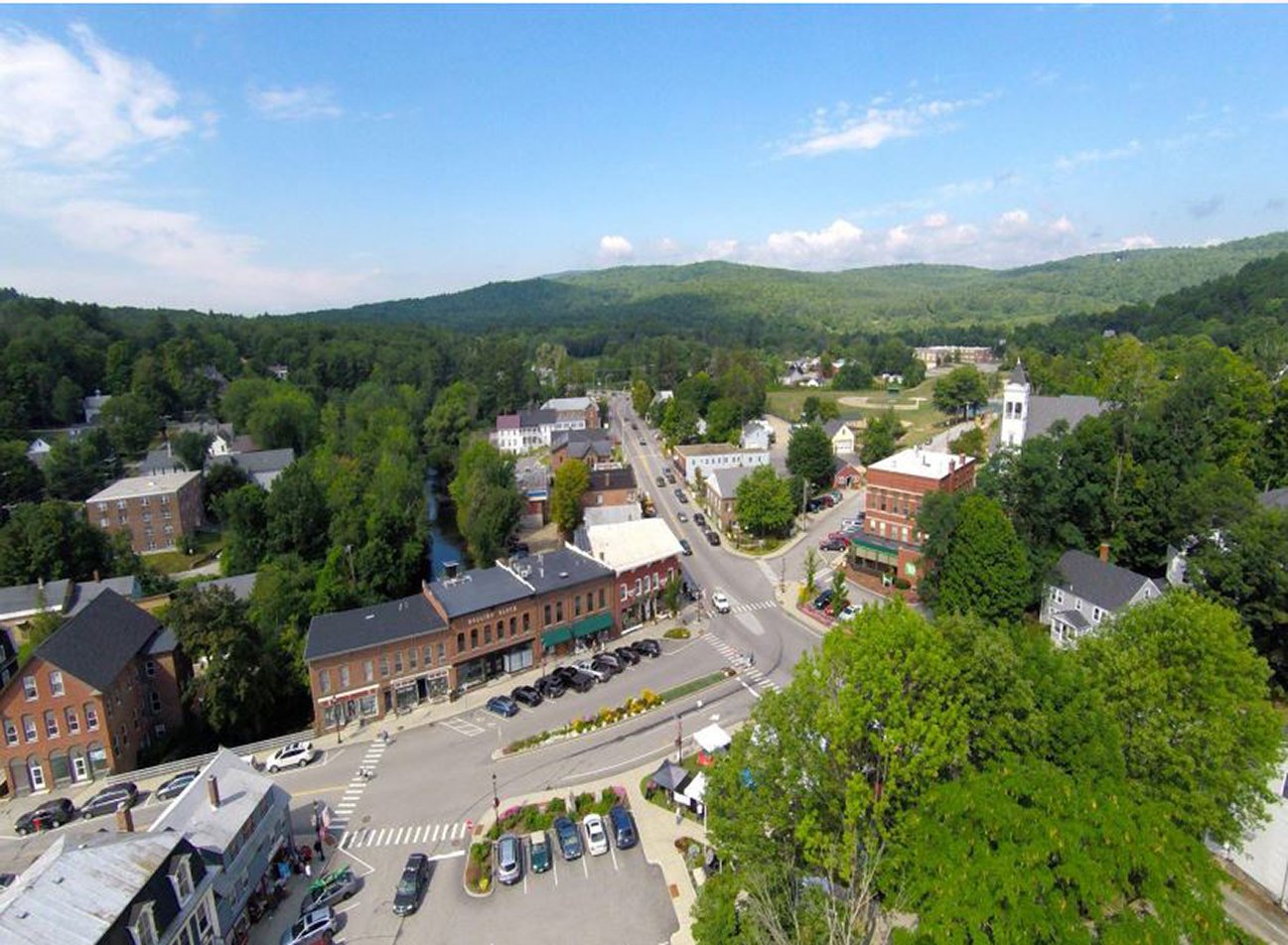 Aerial view of a small town surrounded by green trees and rolling hills under a partly cloudy sky.
