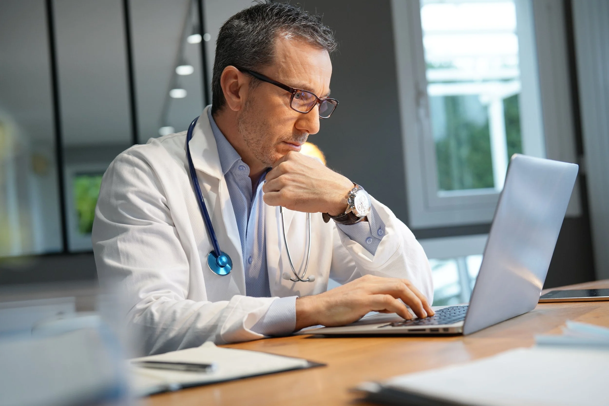 A male doctor wearing a white coat, glasses, and a stethoscope around his neck, working on a laptop at a desk in a modern, well-lit office or medical facility.