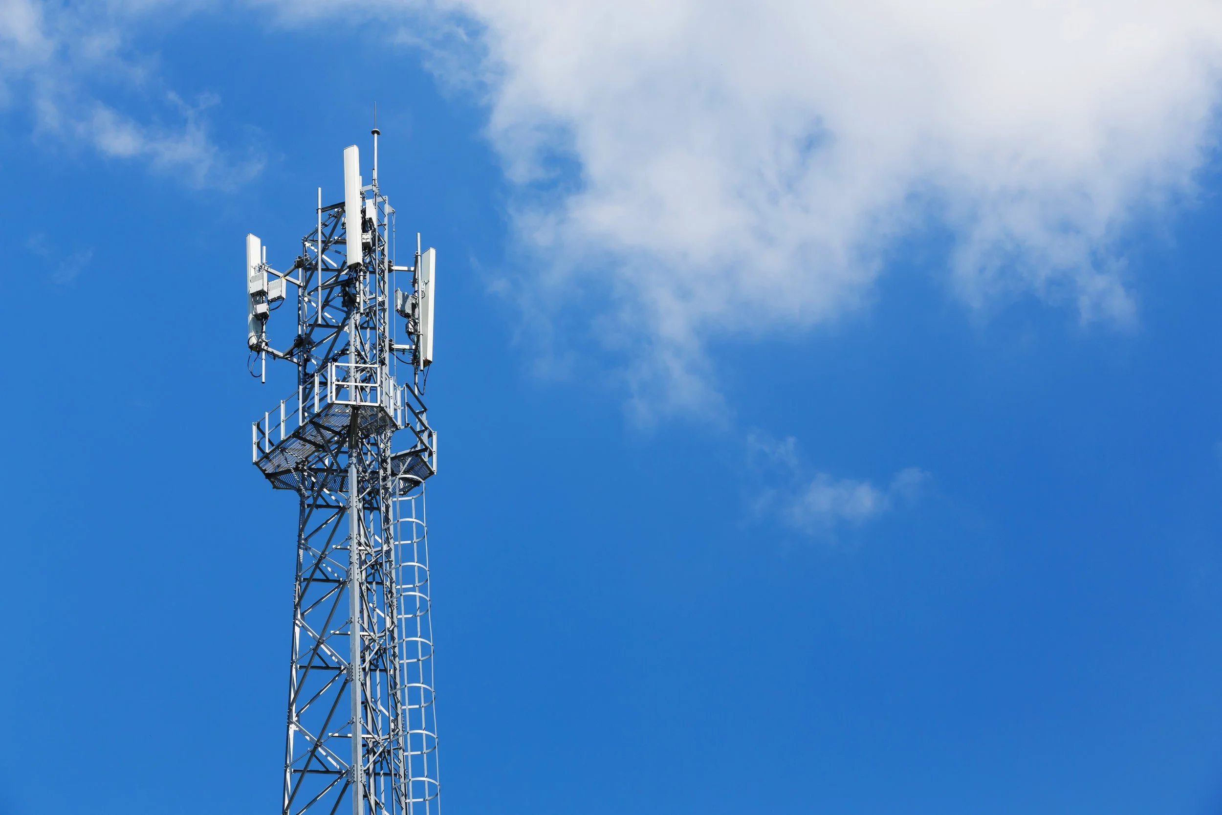 A tall metal communication tower with multiple antennas against a bright blue sky with scattered clouds.
