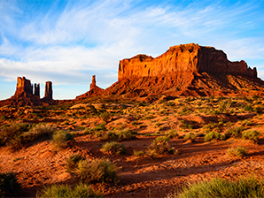 Desert landscape with large rock formations and mesas under a partly cloudy blue sky.