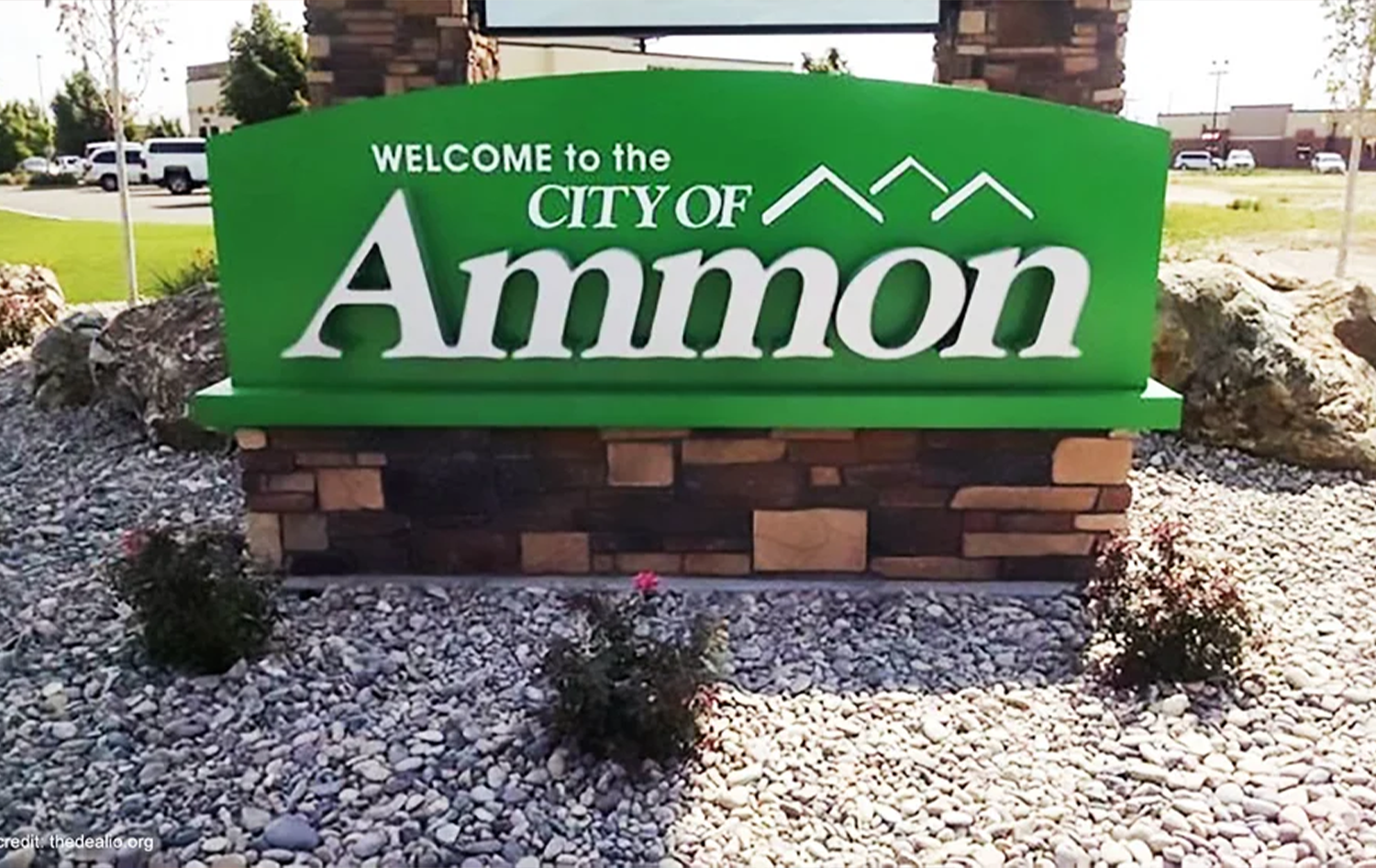 Green welcome sign for the city of Ammon, mounted on a brick base with rocks and small bushes in the foreground.