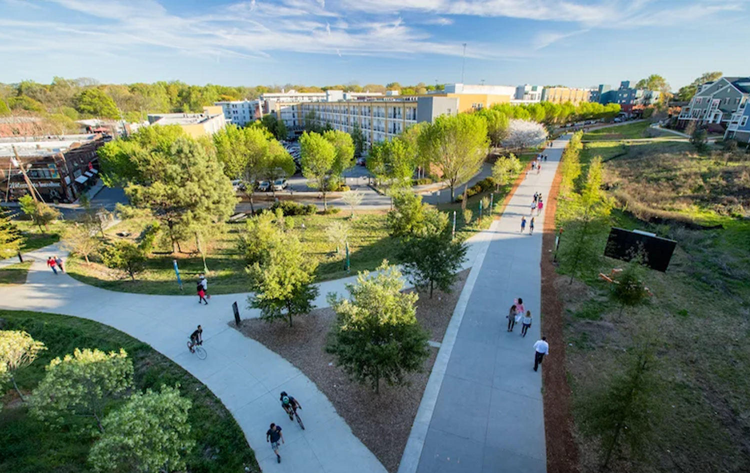 Aerial view of a park with winding pathways, green trees, and people walking and biking, surrounded by modern apartment buildings and commercial area under a blue sky with some clouds.
