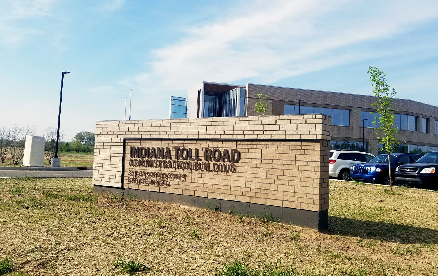 Brick sign for Indiana Toll Road Administration Building, with parking lot and modern office building in background.