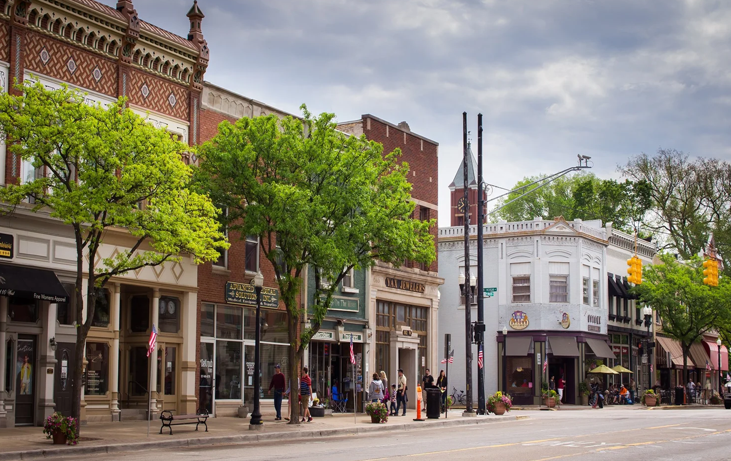 Street view of a small town with historic buildings, trees, and people walking on the sidewalk.