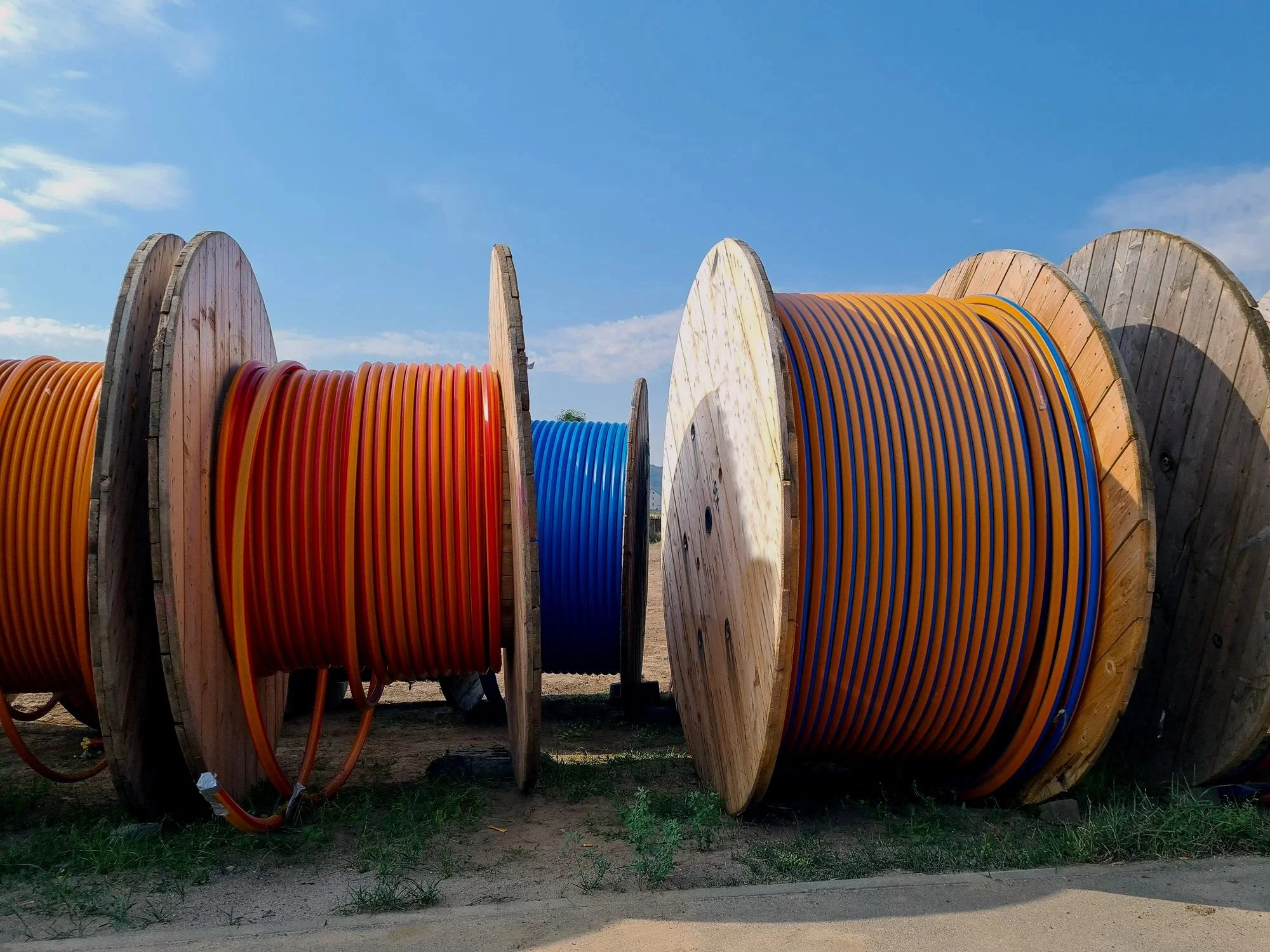 Large wooden spools holding orange, blue, and yellow electrical cables, outdoors under a blue sky.