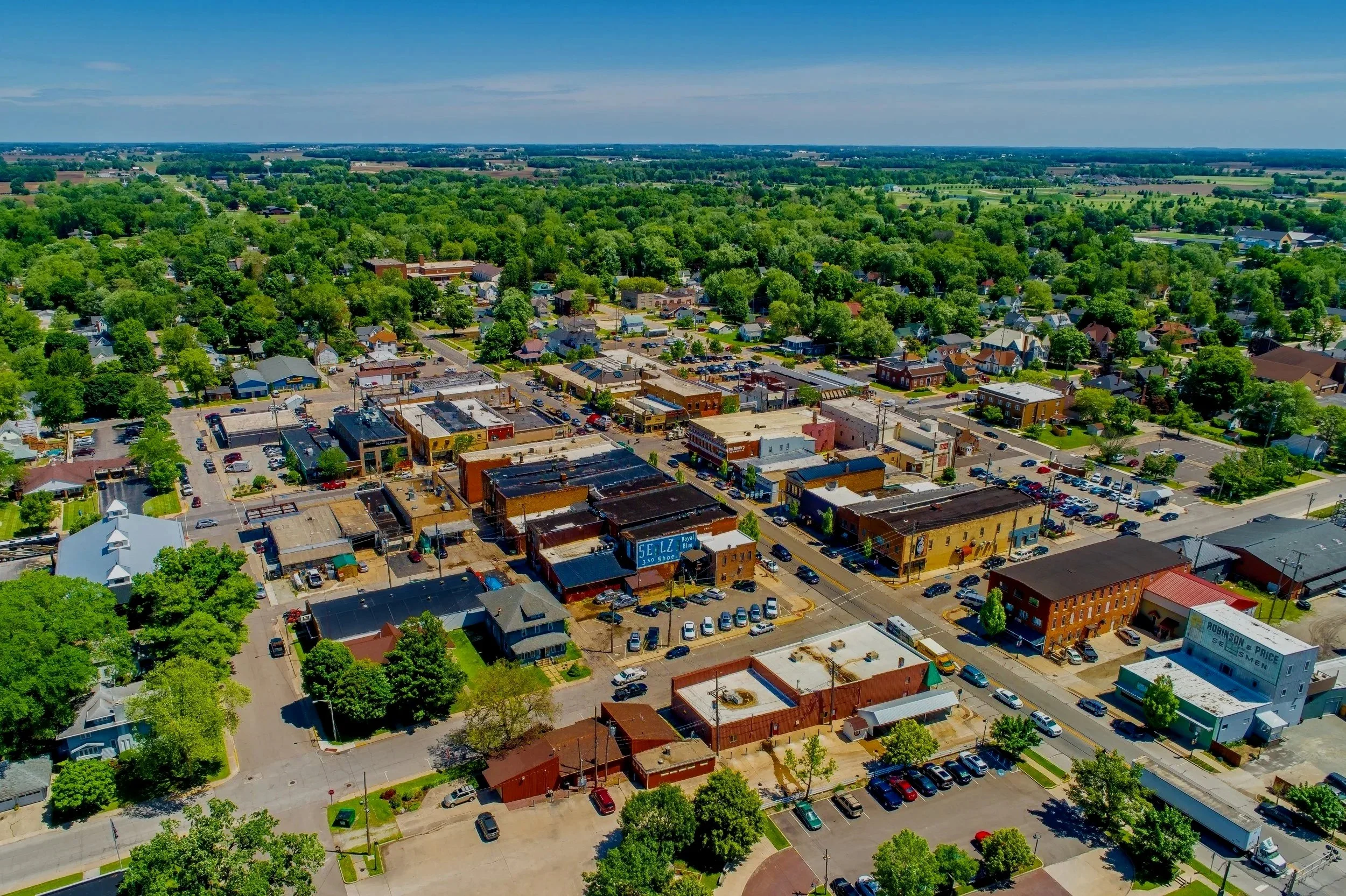 Aerial view of a small town with a commercial district and surrounding green residential neighborhood, green trees, parking lots, and open fields in the distance.
