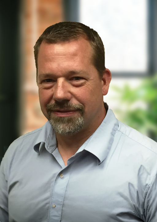 A man with short brown hair and a beard, wearing a light blue button-up shirt, smiling in front of a window with a blurred background of green plants and indoor decor.