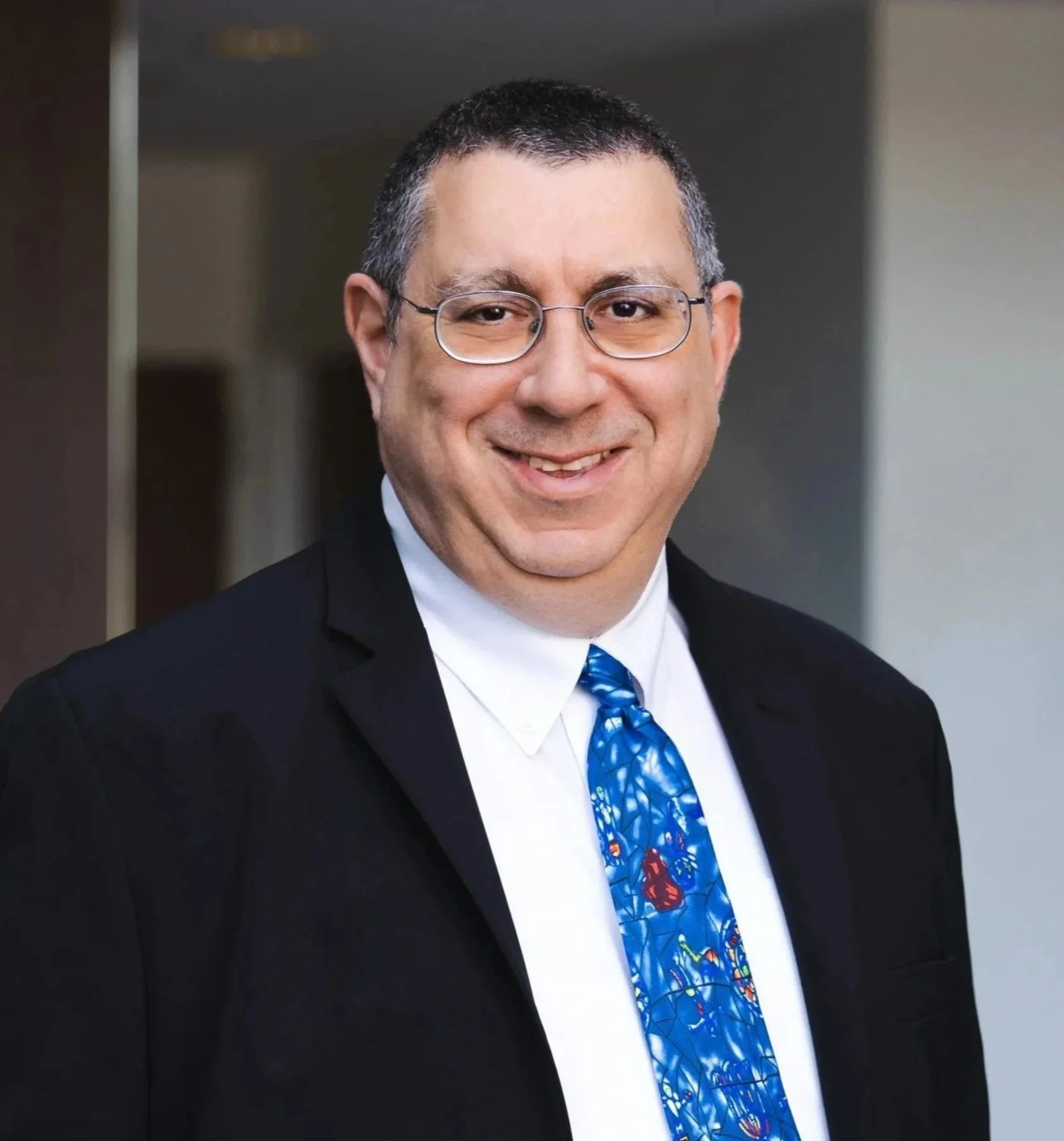 A middle-aged man with short dark hair, wearing glasses, smiling, dressed in a black tuxedo, white shirt, and a colorful tie with a blue background and red and yellow design.