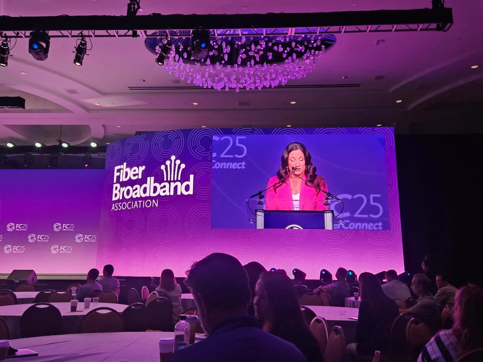 A woman in a pink blazer speaking at a podium during a conference with a large screen behind her displaying the 'Fiber Broadband Association' logo and the text 'FC25 Connect.' The audience is seated at round tables, and the room is decorated with purple lighting and a chandelier.