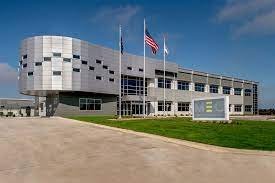 A modern building with an American flag and another flag in front of it, a large lawn, and a blue sky with some clouds.
