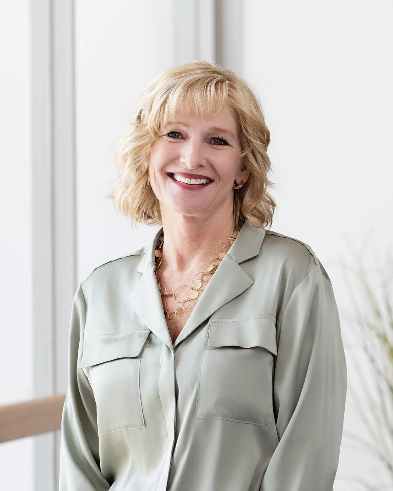 A smiling middle-aged woman with blonde hair wearing a light green blouse and a gold necklace, standing indoors near a window.
