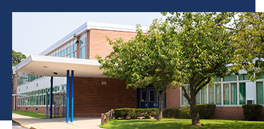 Modern school building with large windows, an overhang entrance, and a landscaped area with a tree in front.
