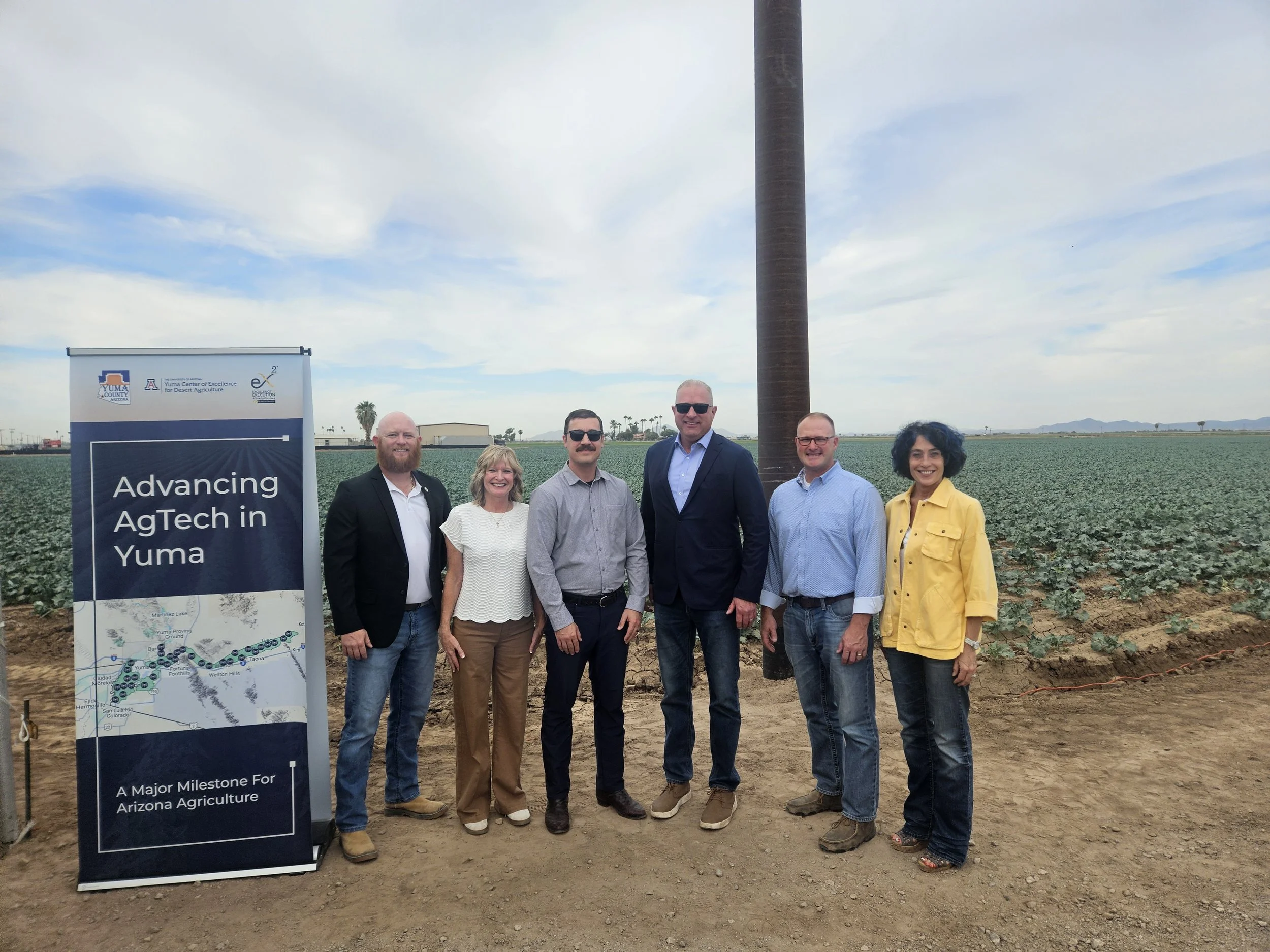 Group of six people standing in a field with a tall structure and a banner that reads 'Advancing Tech in Yuma'. The group includes three men and three women smiling, with a backdrop of farmland and a few distant mountains. The people are dressed in casual and professional attire, and there is a cloudy sky above.