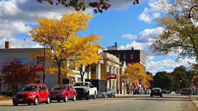 A street lined with trees in fall colors, with yellow and red leaves, and buildings in the background. Several parked cars are visible along the curb under a partly cloudy sky.
