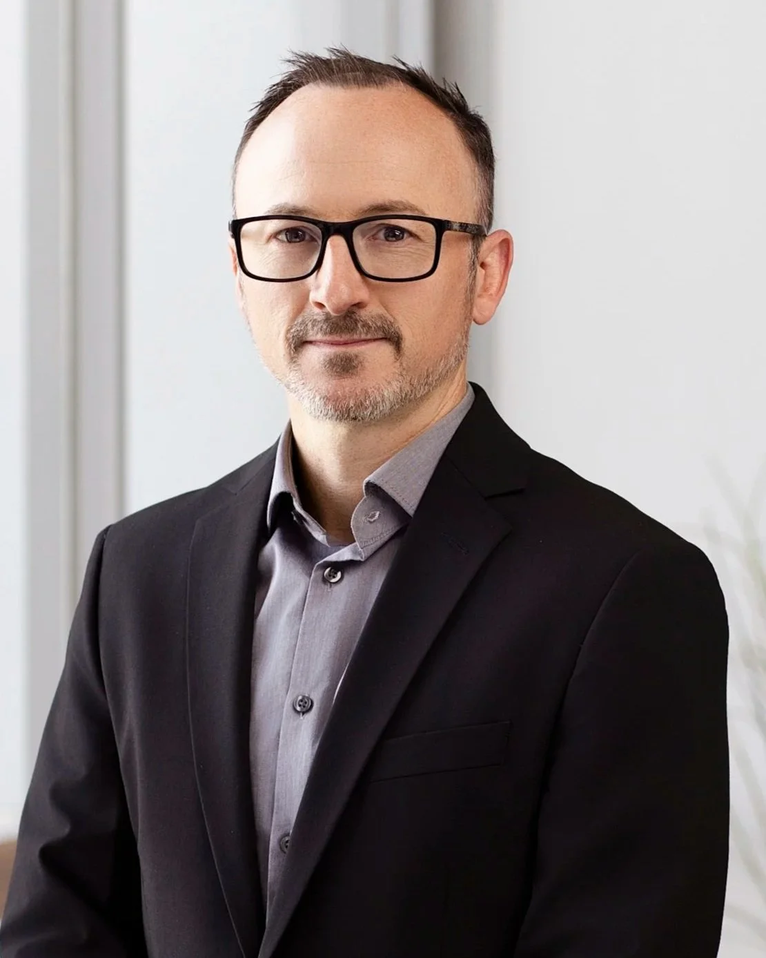 A professional man with glasses, wearing a dark suit and a gray shirt, standing indoors near a window.