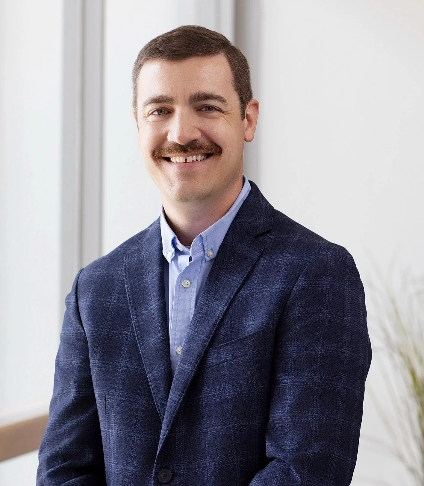 A man with short brown hair, a mustache, and blue eyes, wearing a blue blazer and a light blue button-up shirt, smiling in an indoor setting with a white background.