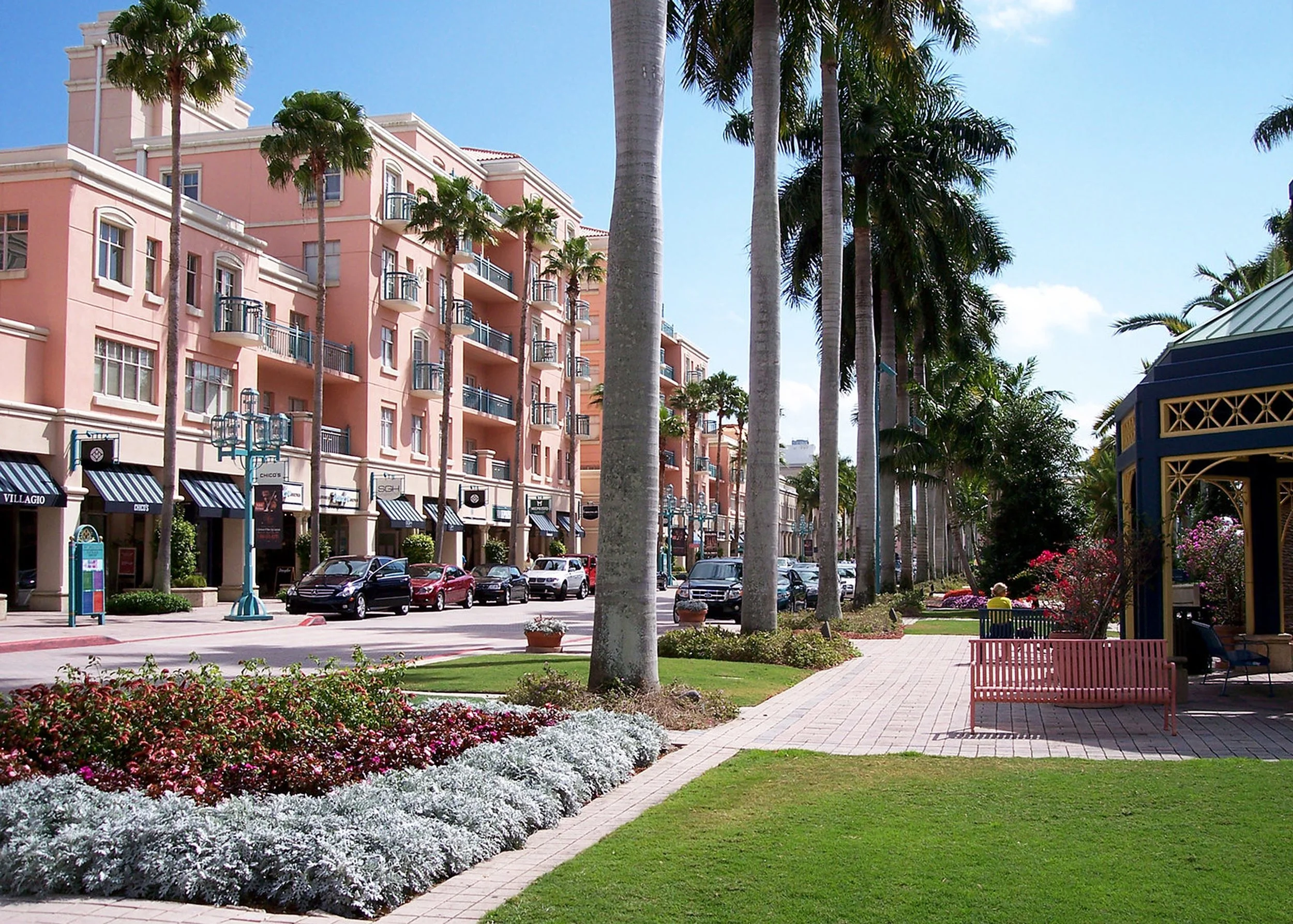 A lively outdoor shopping and dining area under a clear blue sky, featuring a multi-story pink building with balconies, a row of parked cars, and a landscaped garden with colorful flowers, trees, and benches.