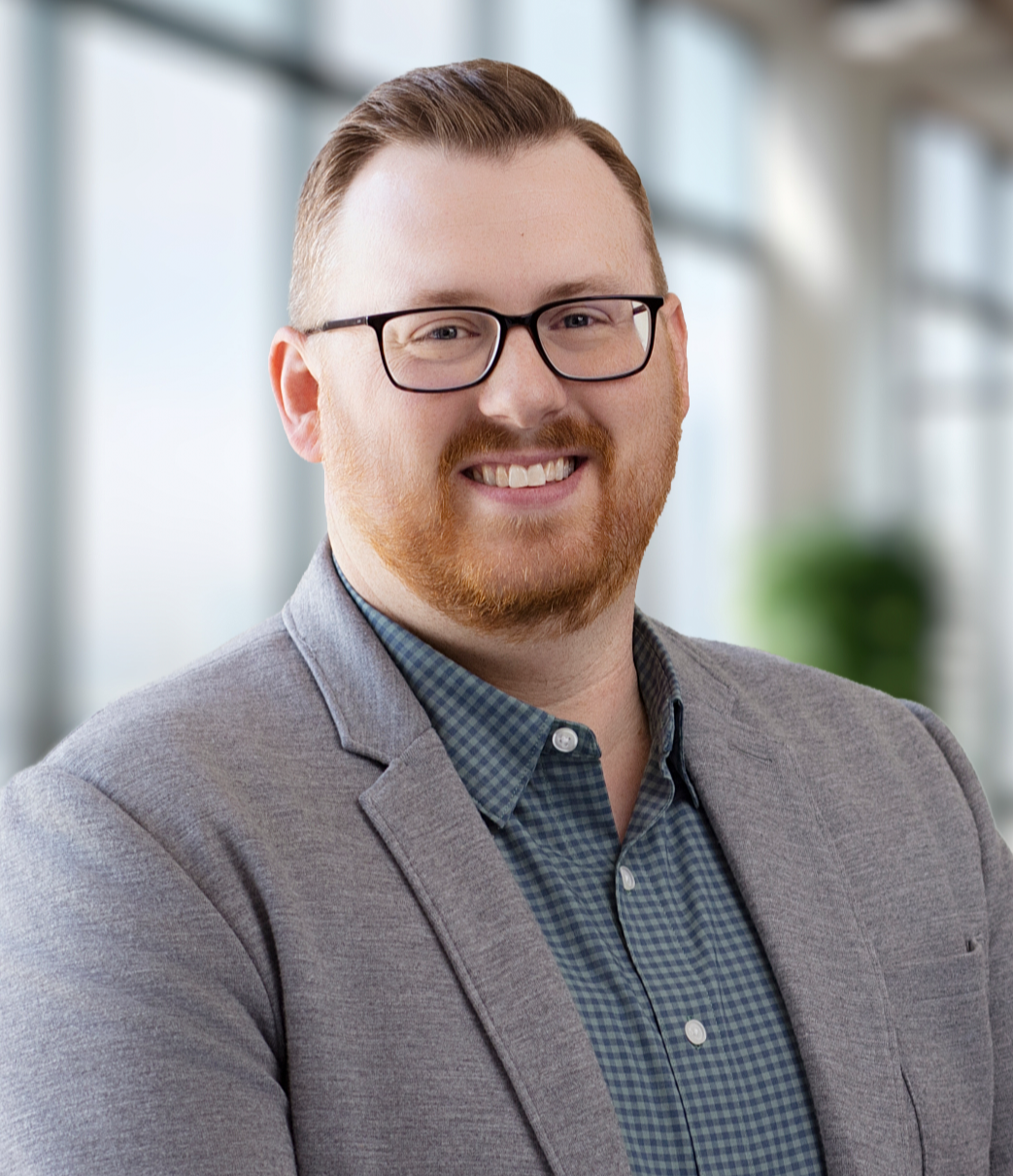 A smiling man with glasses and a red beard, wearing a gray blazer over a blue checkered shirt, standing in a modern office setting with large windows.