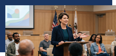 A woman speaks at a podium in a conference room, with an audience of diverse individuals listening attentively, American and California flags in the background.