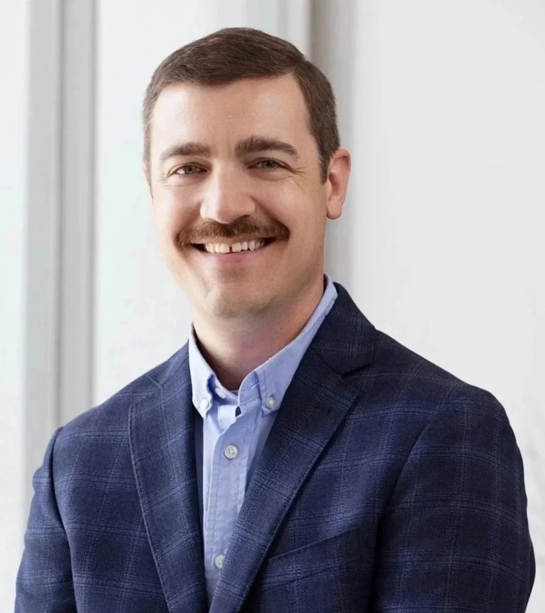 A smiling man with brown hair, a mustache, and a beard, wearing a dark suit jacket and a light blue shirt, standing near a window with a white wall background.