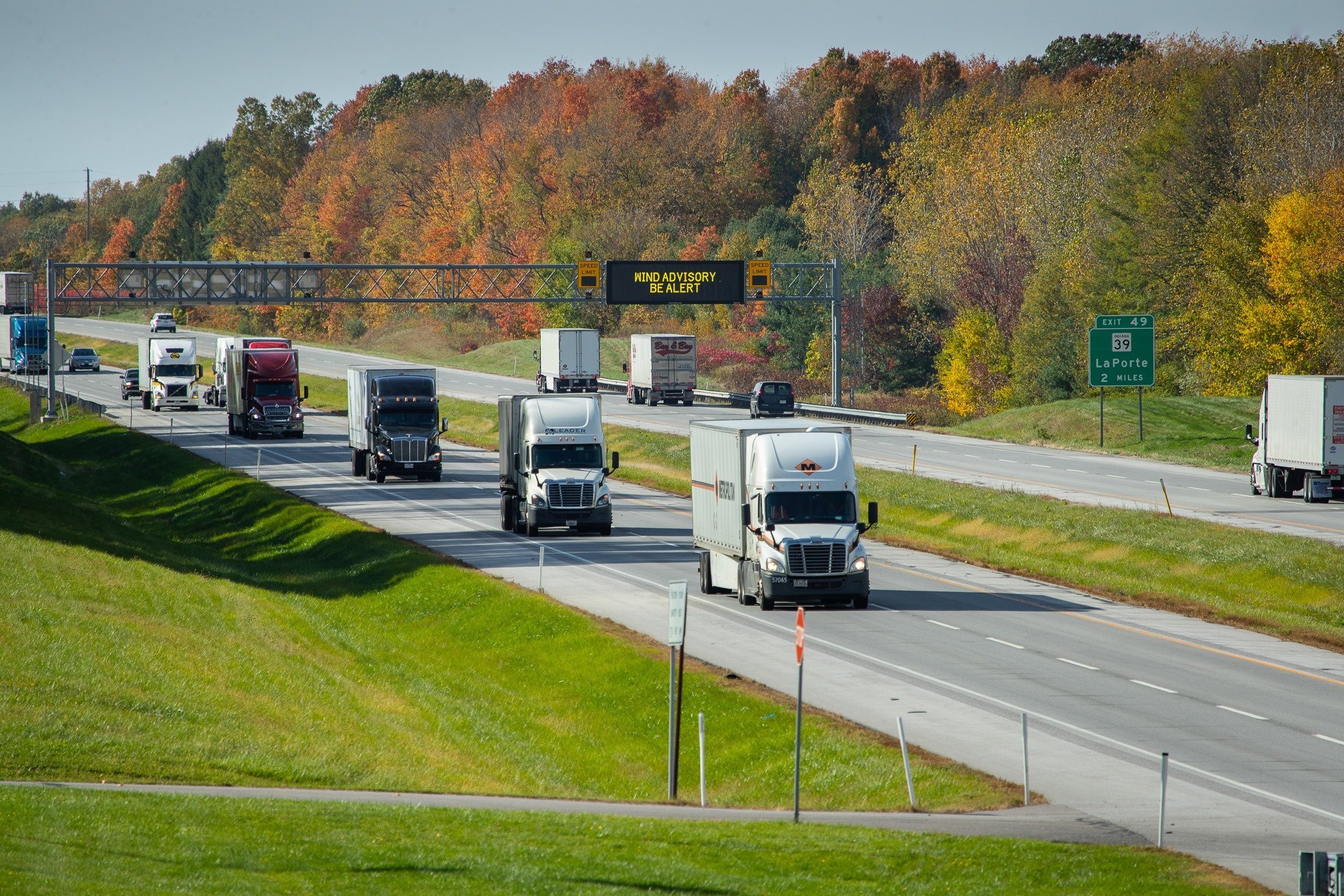 Highway with multiple trucks and cars driving amidst fall foliage, with a digital sign warning about wind advisory.