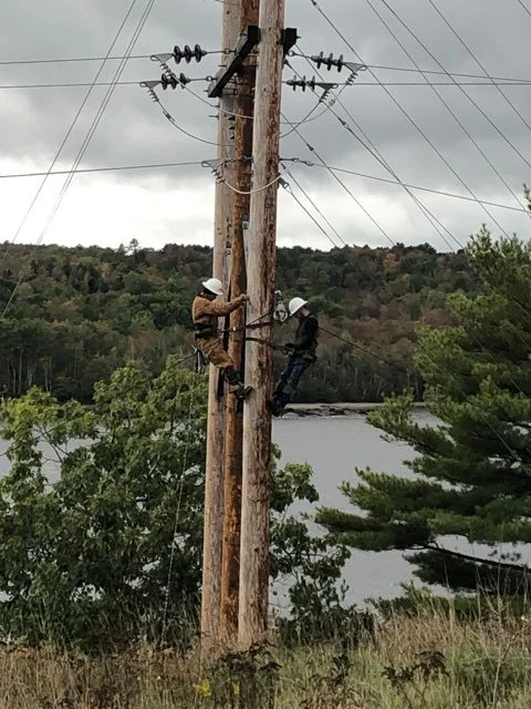 Two workers in safety helmets repairing or inspecting an electric utility pole near a river with forested hills in the background on a cloudy day.