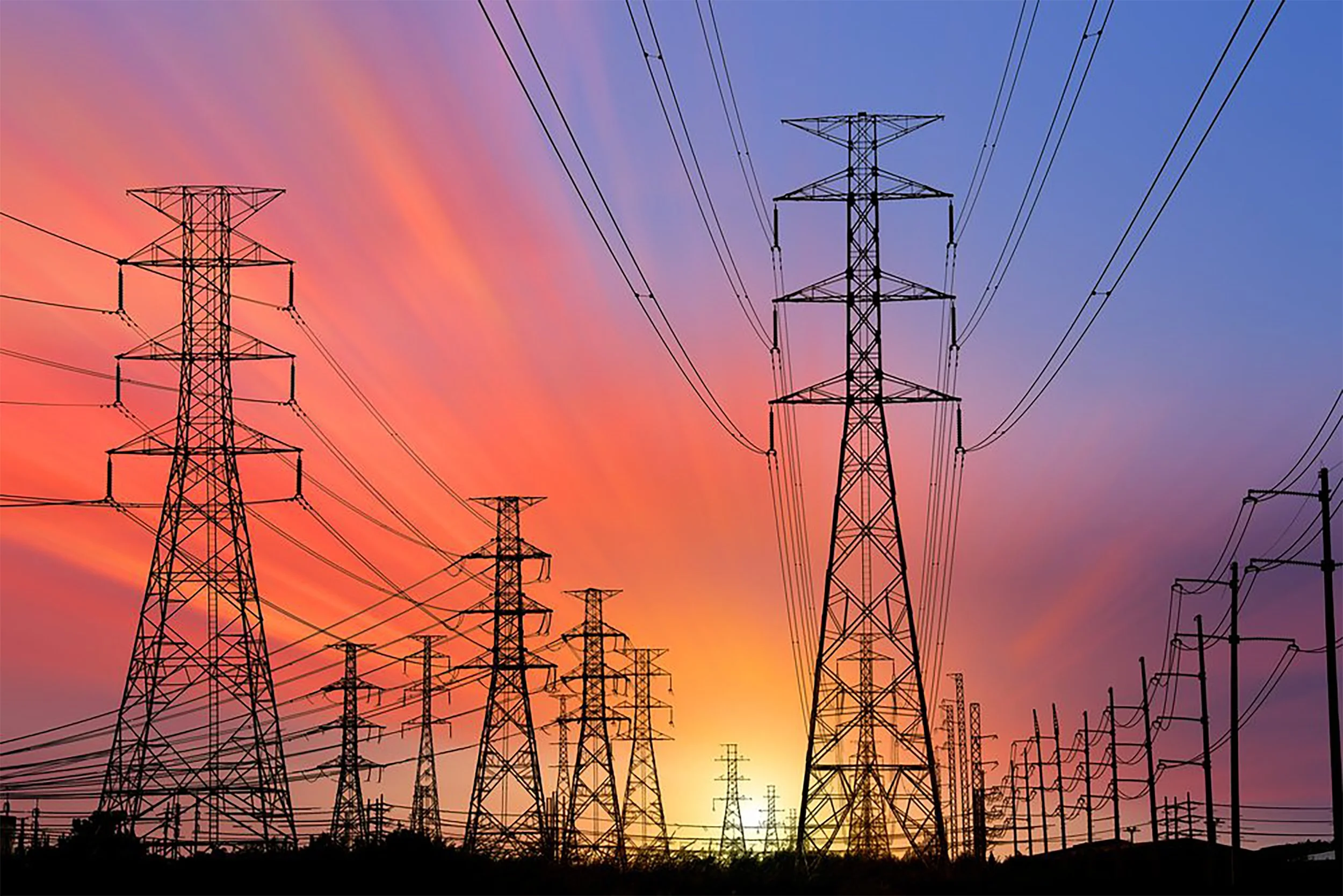 Silhouettes of multiple high-voltage power line towers against a colorful sunset sky with orange, pink, purple, and blue hues.