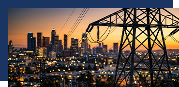 City skyline at sunset with power lines and electrical tower in the foreground.