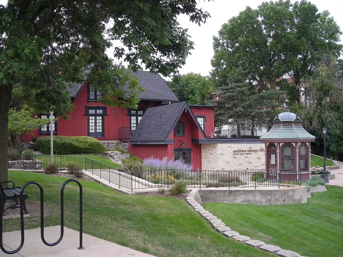 A park with a black metal bike rack, benches, a paved walkway, and a red Victorian-style building with a turret. There is a stone gazebo with a metal roof and a sign reading 'Guthrie Research Center' in front of the building, and trees around the area.