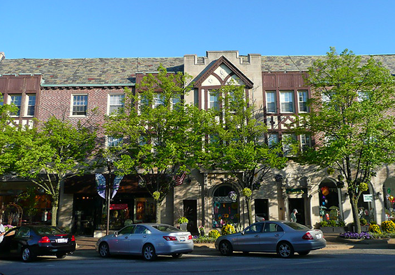 Street view of a building with retail shops on the ground floor and residential or office spaces above, lined with trees and parked cars.