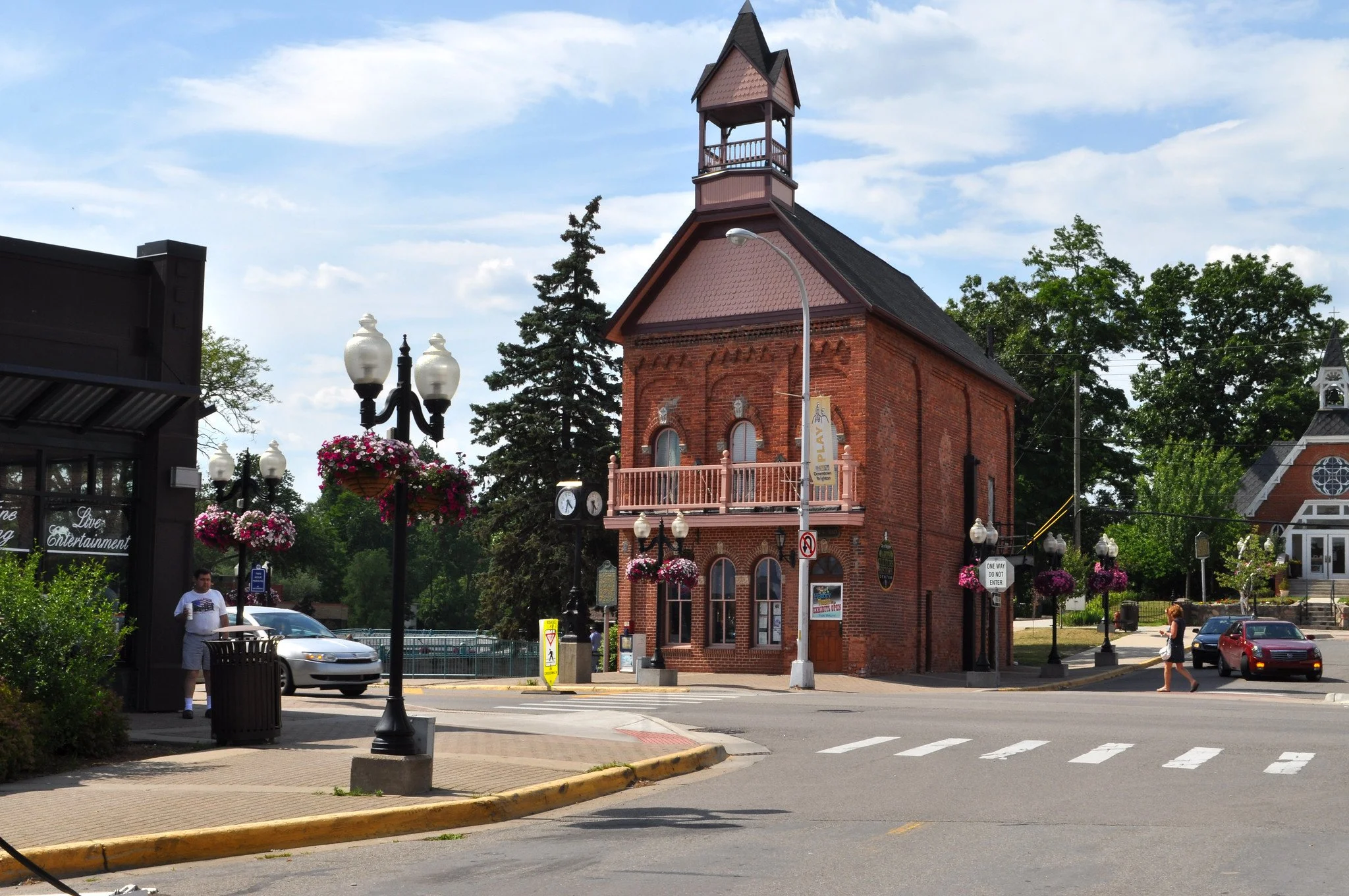 A small red brick building with a clock and a balcony, surrounded by trees, street lamps with hanging flower baskets, and cars in a town square under a partly cloudy sky.