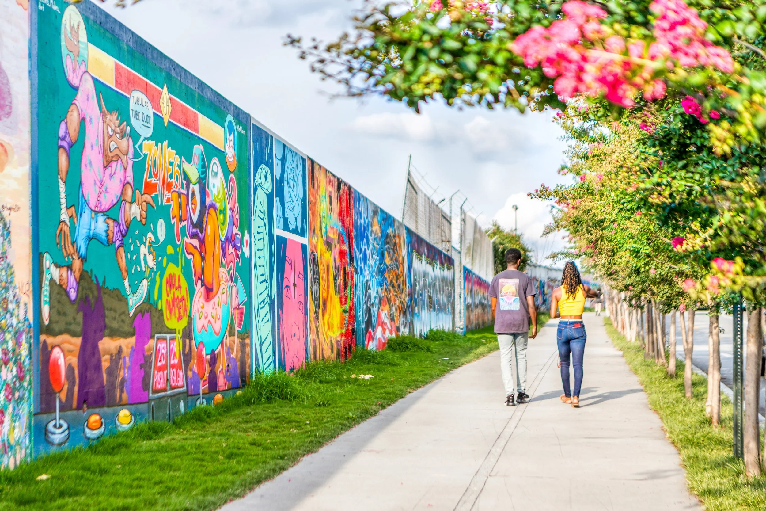 Street view showing a colorful mural on a wall, with two people walking along the sidewalk beside it, and trees with pink flowers lining the street.