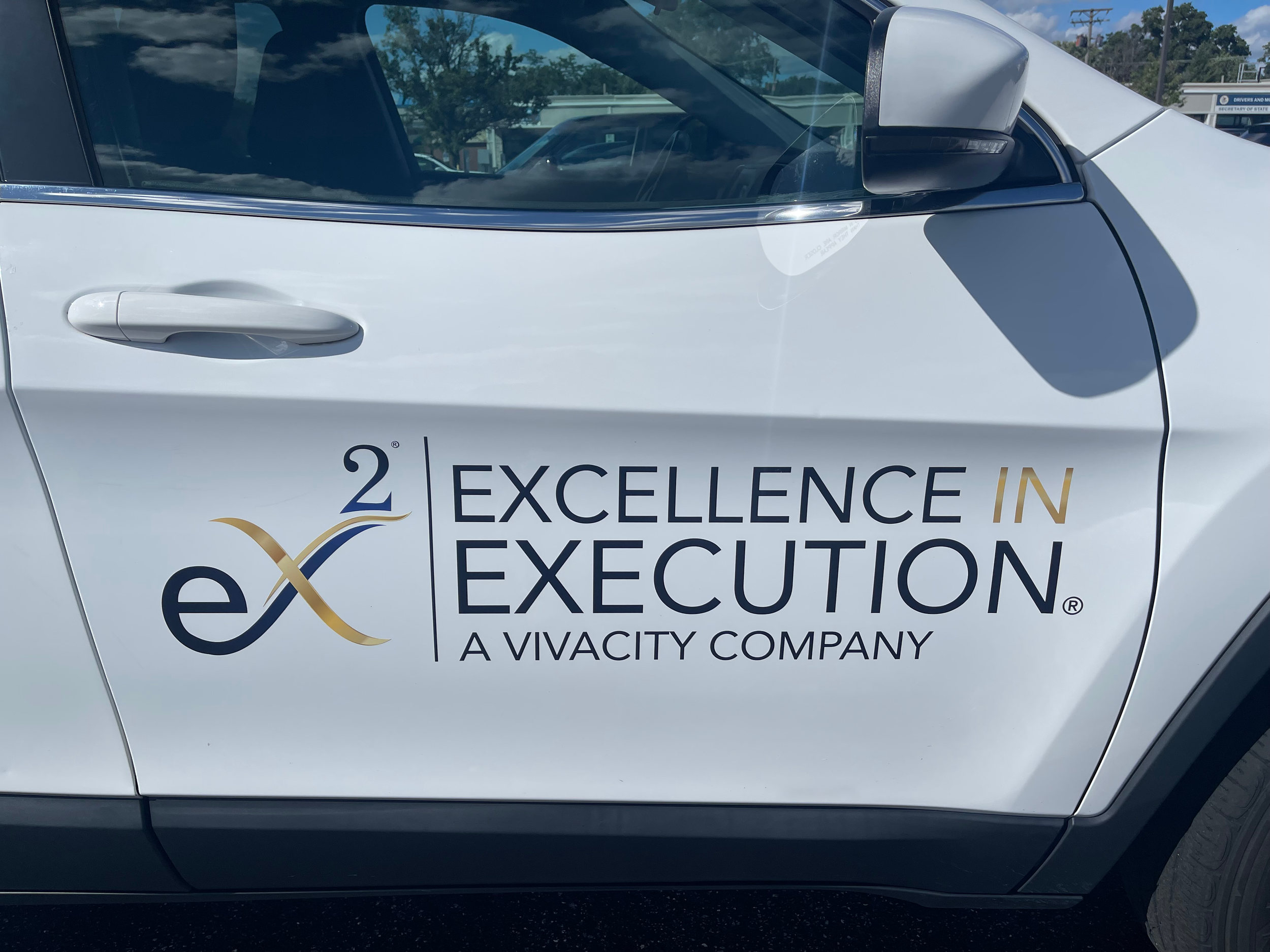 Close-up of a white vehicle with signage for a company called Excelence in Execution, a Vivacity Company, with the vehicle door handle, side mirror, and part of the logo visible.